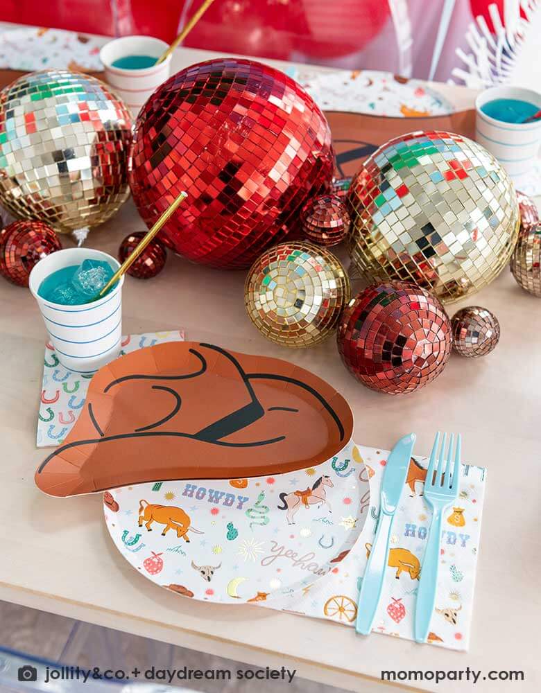 A western cowboy themed party table tablescape featuring Momo Party's cowboy themed tableware by Jollity Co. and Daydream Society including the round cowboy icon patterned round plate, the small napkin, the cowboy hat shaped plate, horseshoe napkin. With disco ball decorations in red and gold in different sizs, making this a modern yet fun tablescape for kid's rodeo, Western or cowboy themed birthday celebration. A perfect inpso for kid's "First Rodeo" first birthday celebration.  