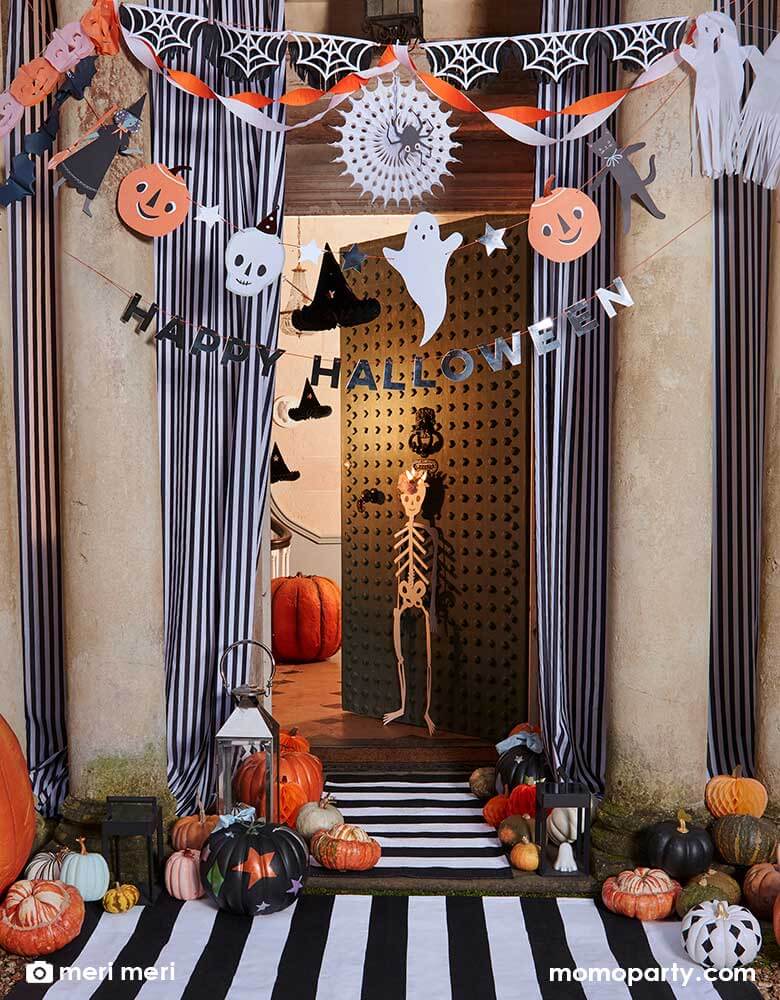 A festive door decorated with a bunch of Halloween decorations and autumn decors including Momo Party's Halloween paper decorations, the This is the Halloween party garland set, skeleton hanging decorations, witch hat honeycombs and various pumpkins in different sizes and colors on the ground. The door is half open as it's inviting all the trick-or-treater to come in for some candies! With black and white striped curtains and doormats, this makes a great inspo for Halloween home porch decorations.