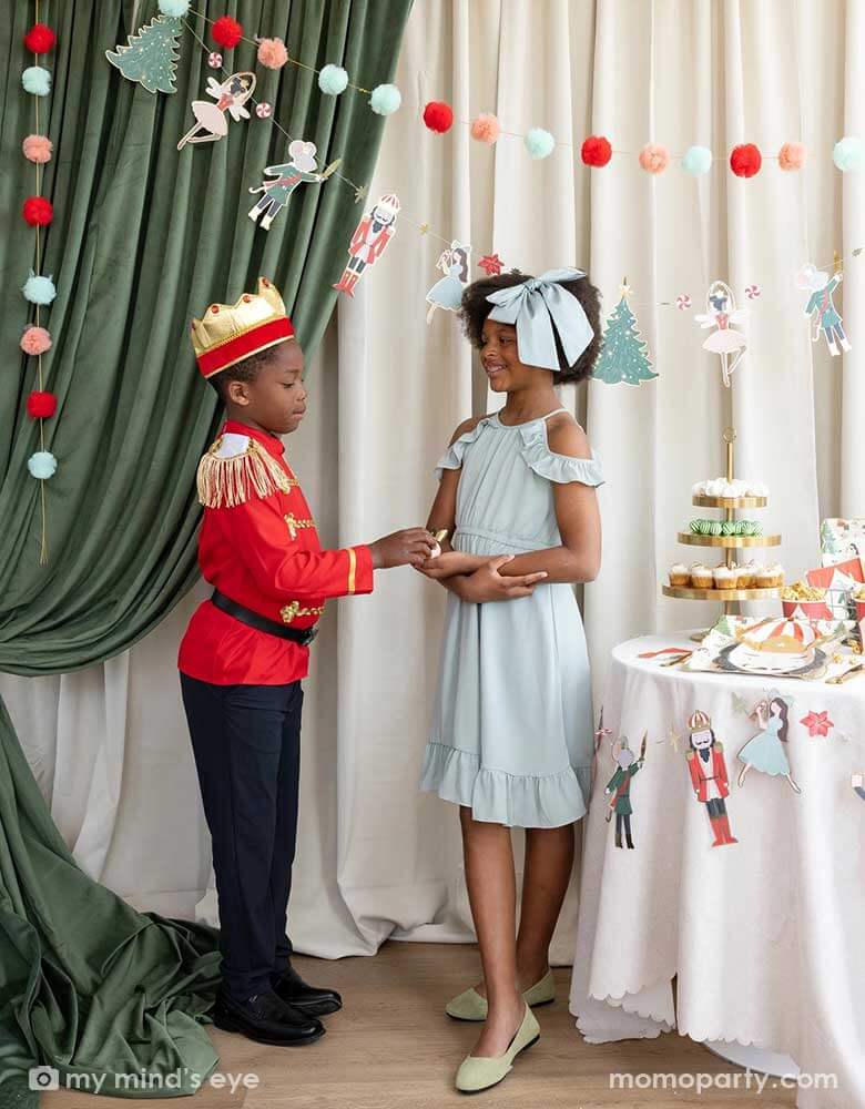 A girl and a boy dressed as the Nutcracker characters in a Nutcracker themed holiday party. They're standing next to a party table filled with Holiday treats and Nutcracker themed party supplies. In their back there's a stage curtain in satin green and beige which is decorated with Momo Party's Nutcracker 6' garland with pom-poms which adds a festive touch to the Holiday decoration.