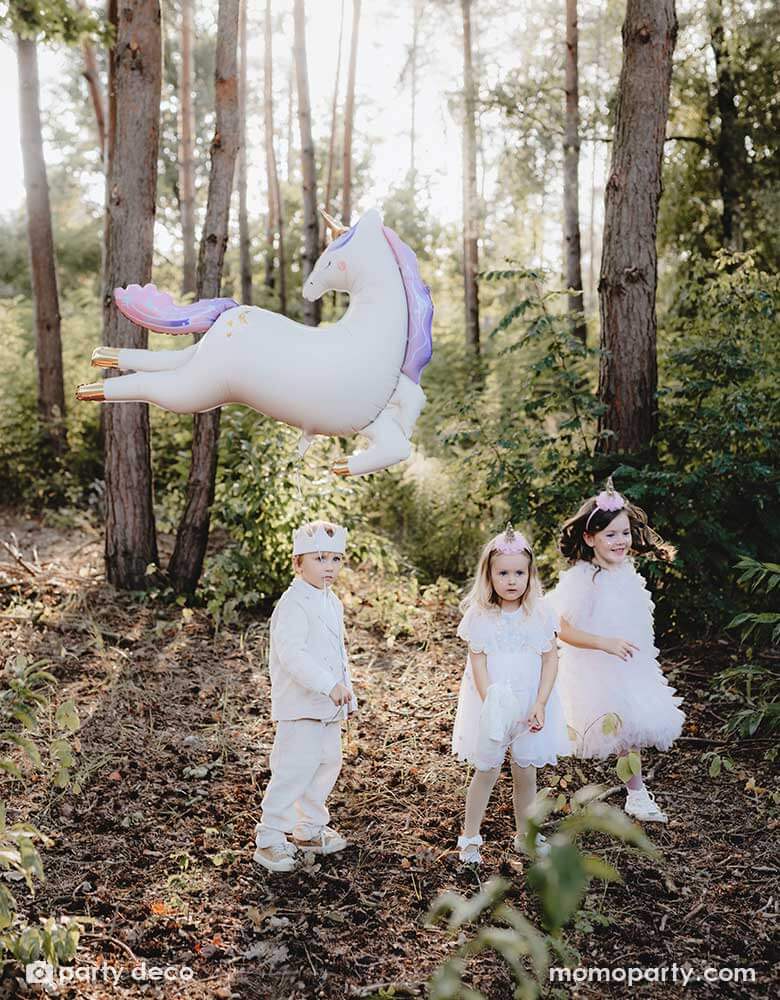 Three kids dressed in white clothing with party hats in an enchanting forest with Momo Party's enchanting unicorn shaped foil balloon floating in the air, making this a dreamy scene for kid's special celebration.