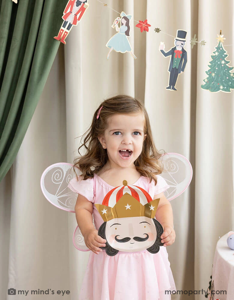 A little girl dressed up as Sugar Plum Fairy from Nutcracker's holding Momo Party's 10" Nutcracker head shaped party plate. In her back there's a stage curtain in satin green and beige which is decorated with Momo Party's Nutcracker garland with pom-poms which adds a festive touch to the Holiday decoration.