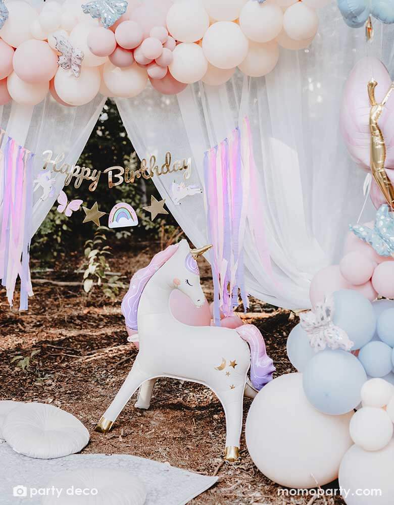 An enchanting unicorn themed birthday party backdrop decoration featuring Momo Party's 37.5" x 34" unicorn standing foil balloon around beautiful pastel colored balloon garlands adorned with butterfly foil balloons and a gold Happy Birthday banner with pastel streamers in the front of a dreamy curtain, making this a whimsical scene for a girly's whimsical unicorn themed birthday party.