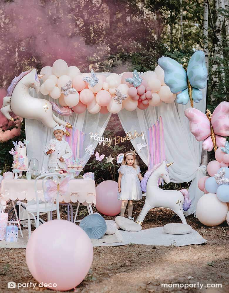 Two kids dressed up as fairies standing in an enchanting unicorn themed birthday party backdrop decoration featuring Momo Party's 37.5" x 34" unicorn standing foil balloon around beautiful pastel colored balloon garlands adorned with butterfly foil balloons and a gold Happy Birthday banner with pastel streamers in the front of a dreamy curtain. In the front there's a party table with a two tier birthday cake and cupcakes, making this a whimsical scene for a girly's whimsical unicorn themed birthday party.