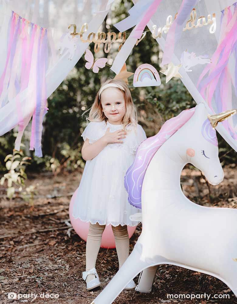 A girl in a white dress standing in an enchanting unicorn themed birthday party backdrop featuring Momo Party's 37.5" x 34" unicorn standing foil balloon around a gold Happy Birthday banner with pastel streamers hung in the front of a dreamy curtain, making this a whimsical scene for a girly's whimsical unicorn themed birthday party.