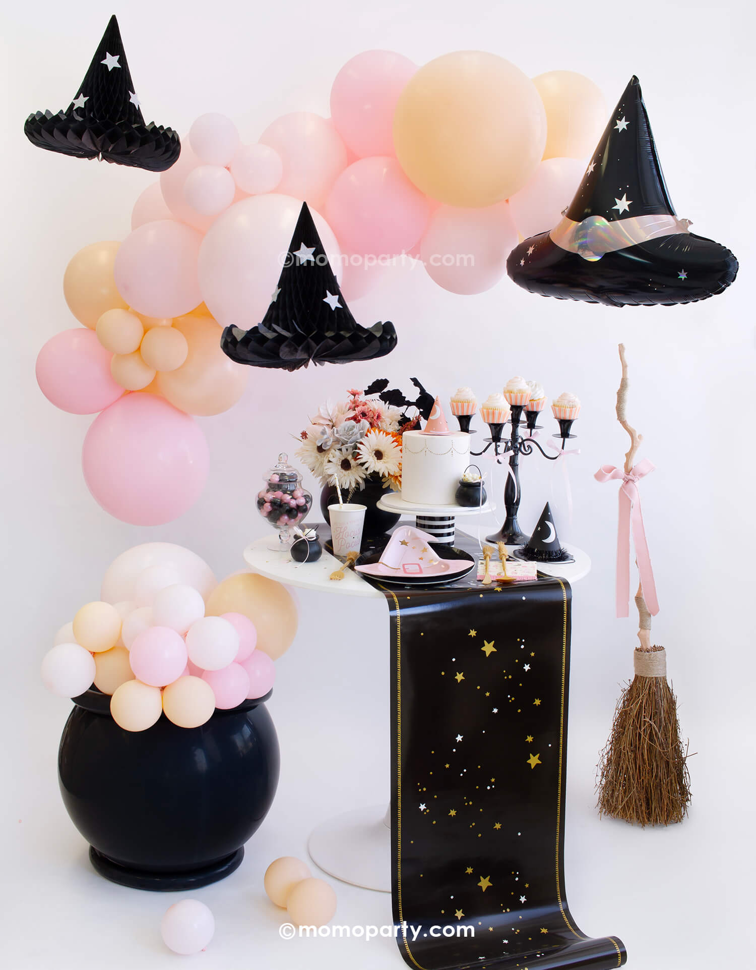 Momo-Party 2024 Halloween Party - Hocus Pocus Pink theme with hanging honeycomb witch hats Decorations and pink witch hat foil balloon from the ceiling. Featuring a Pocus Pink Halloween balloon garland, DIY cauldron balloon decor, and a decorative witch broom with a pink bow. Witch-themed tableware includes Onyx Black dinner plates, pink witch hat-shaped plates, Hocus Pocus Pink fringe napkins,and cups, Halloween witch's broom cutlery on a Mystical Stars table runner. pink witch themed birthday party