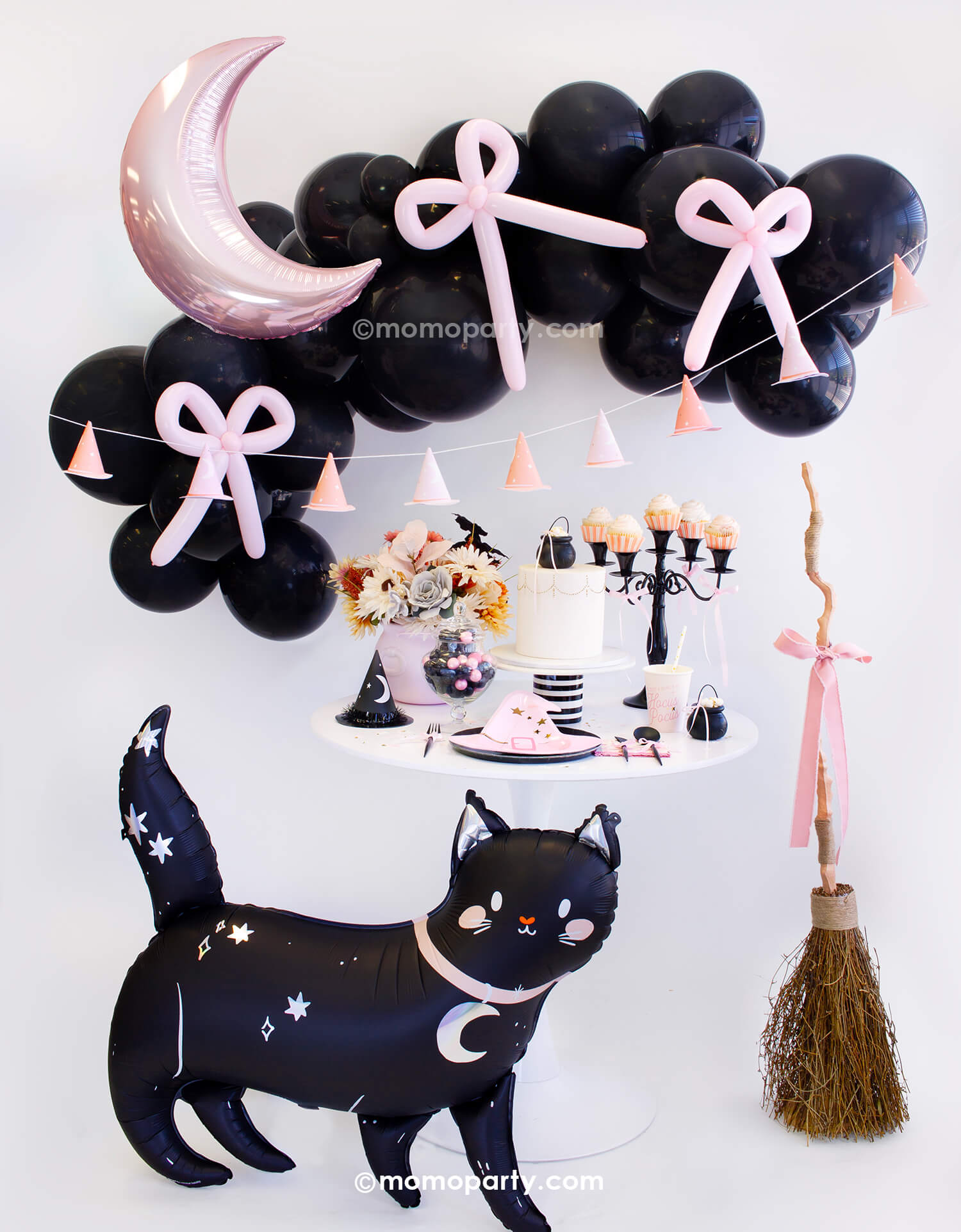 An adorable kids friendly witch themed Halloween party look styled by Momo Party. With black balloon garland adorned with pastel pink balloon bows and the pastel pink crescent moon shaped foil balloon, along with pink witch hat party banner as the backdrop. On the party table there are witch themed tableware including pink witch hat shaped plates, Hocus Pocus napkins and party cups, along with mini cauldrons and witch hats. In the front there are a friendly black cat foil balloon and a pink bow broom.