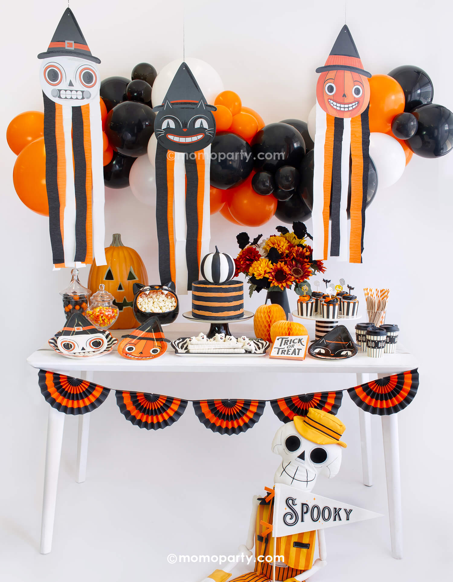 Momo Party's vintage Halloween party box displayed with classic themed decorations including black and orange hanging decorations with streamers, vintage-style tableware including pumpkin, skull and black cat shaped plates, striped party cups and trick or treat napkins, with a orange and black bunting garland and fun centerpieces like jack-o-lantern decoration and fall flower arrangement, it's perfect for a nostalgic Halloween celebration this spooky season.