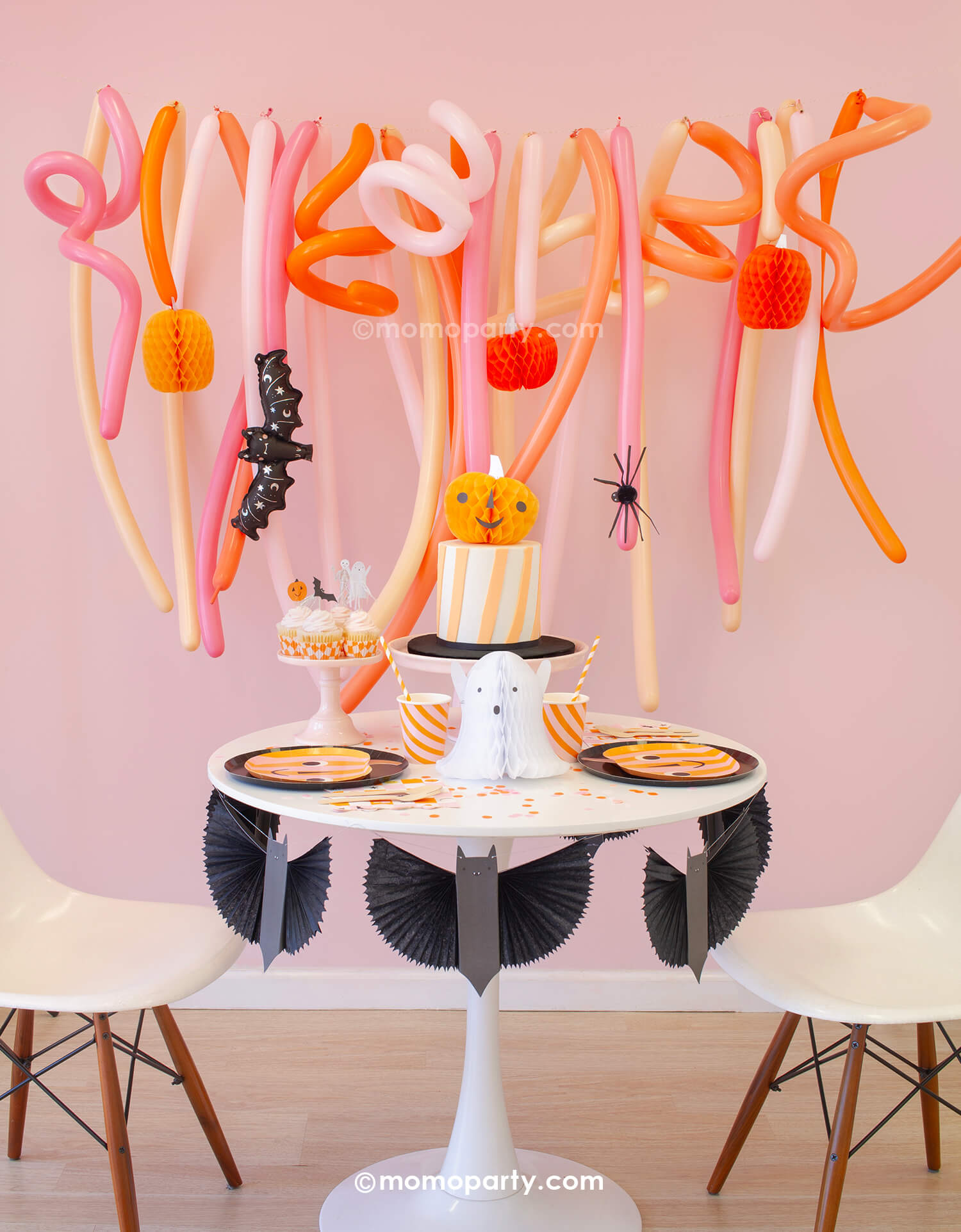 Momo Party's Trick or Treat Pink Halloween box set up featuring a festive long balloon backdrop with curling long balloons  in coral, pink, rose and peach colors. On the balloon backdrop it's decorated with paper pumpkin honeycombs and a mini bat foil balloon. In front of the balloons, a small round table is filled with Halloween party supplies including pumpkin shaped plates, a bat garland, ghost and pumpkin honeycomb decorations from momoparty.com.