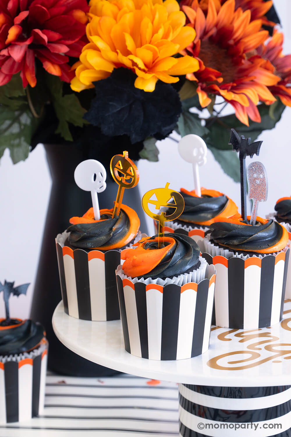 A close-up shot of Halloween orange and black swirled cupcakes in Momo Party's vintage Halloween black striped food cups topped with monster mash acrylic toppers featuring Halloween characters like pumpkin, skull, bat and ghost. The cupcakes are sitting on a black striped cupcake holder which is placed on a black striped table runner. On the table there's a fall flower arrangement with creepy black leaves, making this a perfect inspo for a fun and festive Halloween bash in this spooky season.