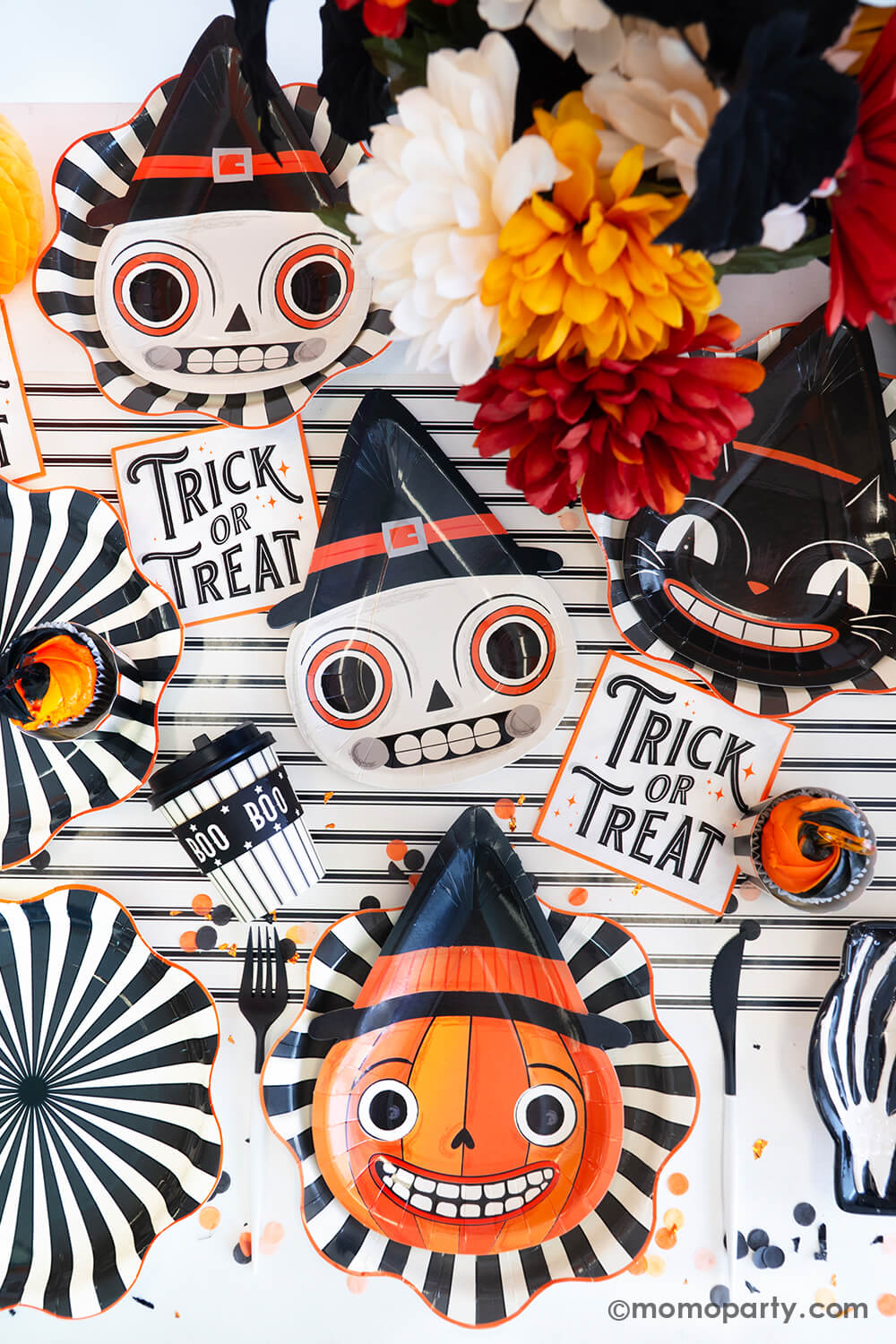 A fun and festive Halloween party table featuring Momo Party's vintage Halloween tableware including skull, pumpkin and black cat shaped party plates on top of the black striped starburst dinner plates, with trick or treat small napkins and BOO to-go cozy cups on top of the cream and black striped table runner, with a sprinkle of orange and black confetti and fall flower arrangement, this provides a perfect inpo for a nostalgic Halloween celebration this spooky season!