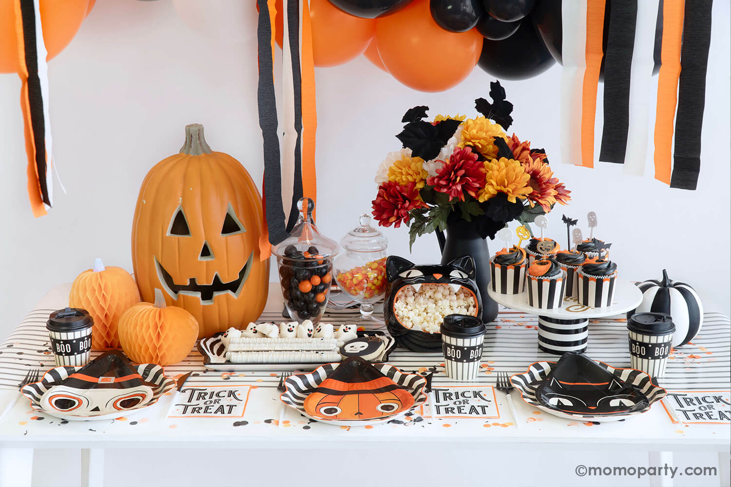 A vintage Halloween party table featuring various party supplies from Momo Party including black striped starburst dinner plates, vintage Halloween character shaped side plates including the skull, the pumpkin and the black cat, Boo! to-go party cups, trick or treat small napkins, black striped food cups, honeycomb pumpkin decorations and monster mash acrylic cupcake toppers. With Halloween treats and fall flower arrangement, this makes a fun and festive kid's friendly Halloween bash in this spooky season!