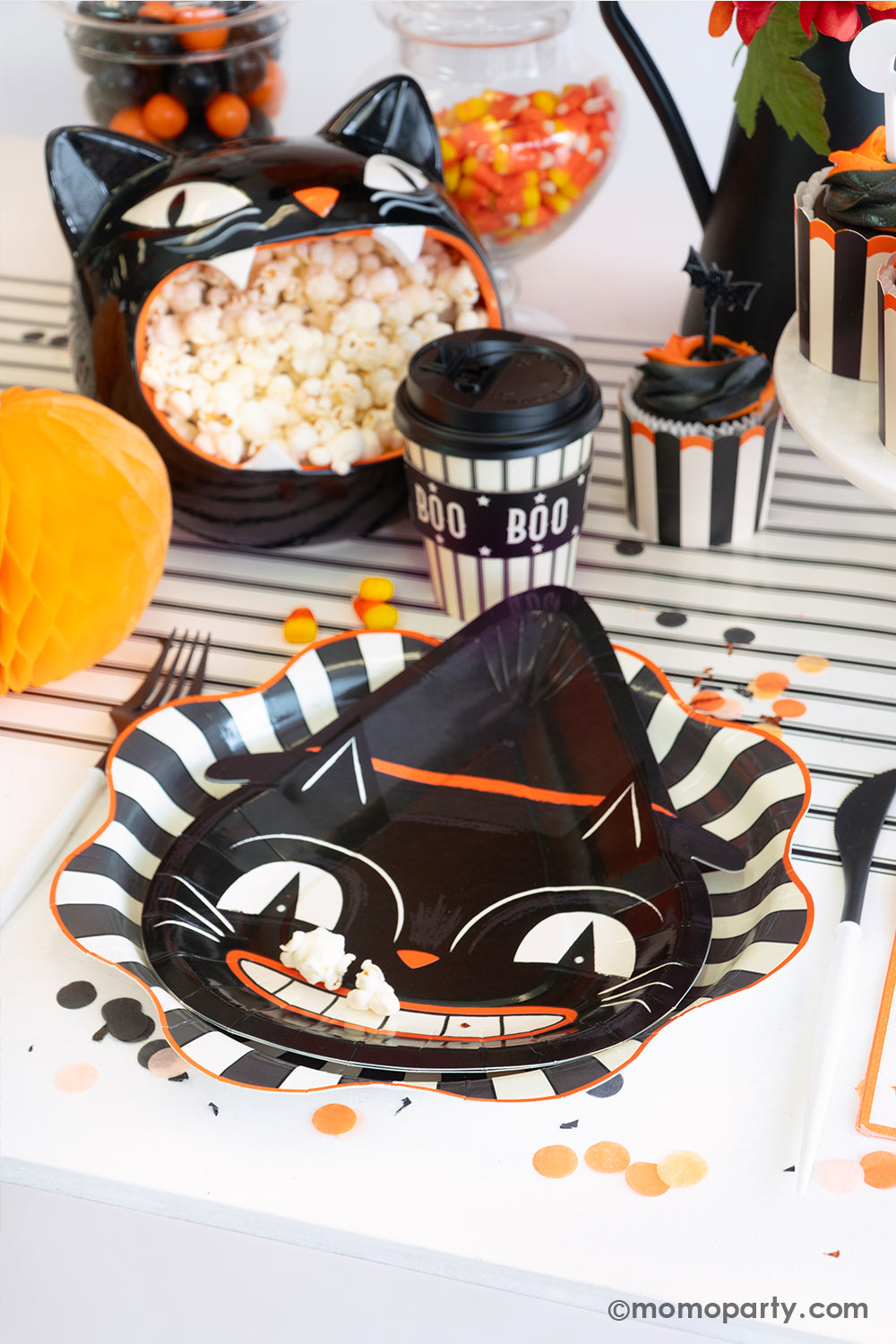 A fun and festive Halloween place setting featuring Momo Party's vintage Halloween party supplies including the black striped starburst plate layered with the black cat shaped side plates. With the trick or treat small napkins, Boo! to-go party cups and black striped food cups filled with cupcakes on the modern black and cream striped  table runner. On the table there are various treats, candies and popcorn, making this an eerie yet fun table setting idea for a festive Halloween celebration for kids.