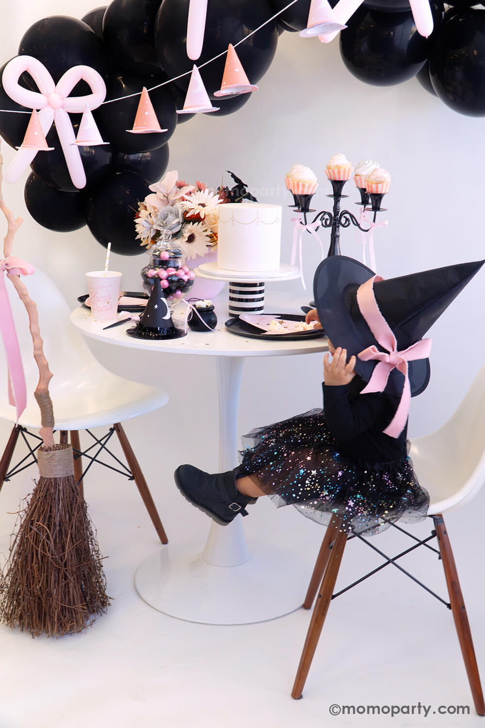 A little girl dressed as Halloween witch with an adorable pink bow on her witch hat setting at a cute witch-inspired Halloween party table featuring various pink Halloween party supplies from Momo Party including pink witch hat shaped plates, Hocus Pocus pink party cups, Hocus Pocus pink small napkins, with a pink floral arrangement and a black candle holder holding pink cupcakes on the table. In the back there's a black balloon garland adorned with pink bow balloons, with a broom adorned with a pink bow. 