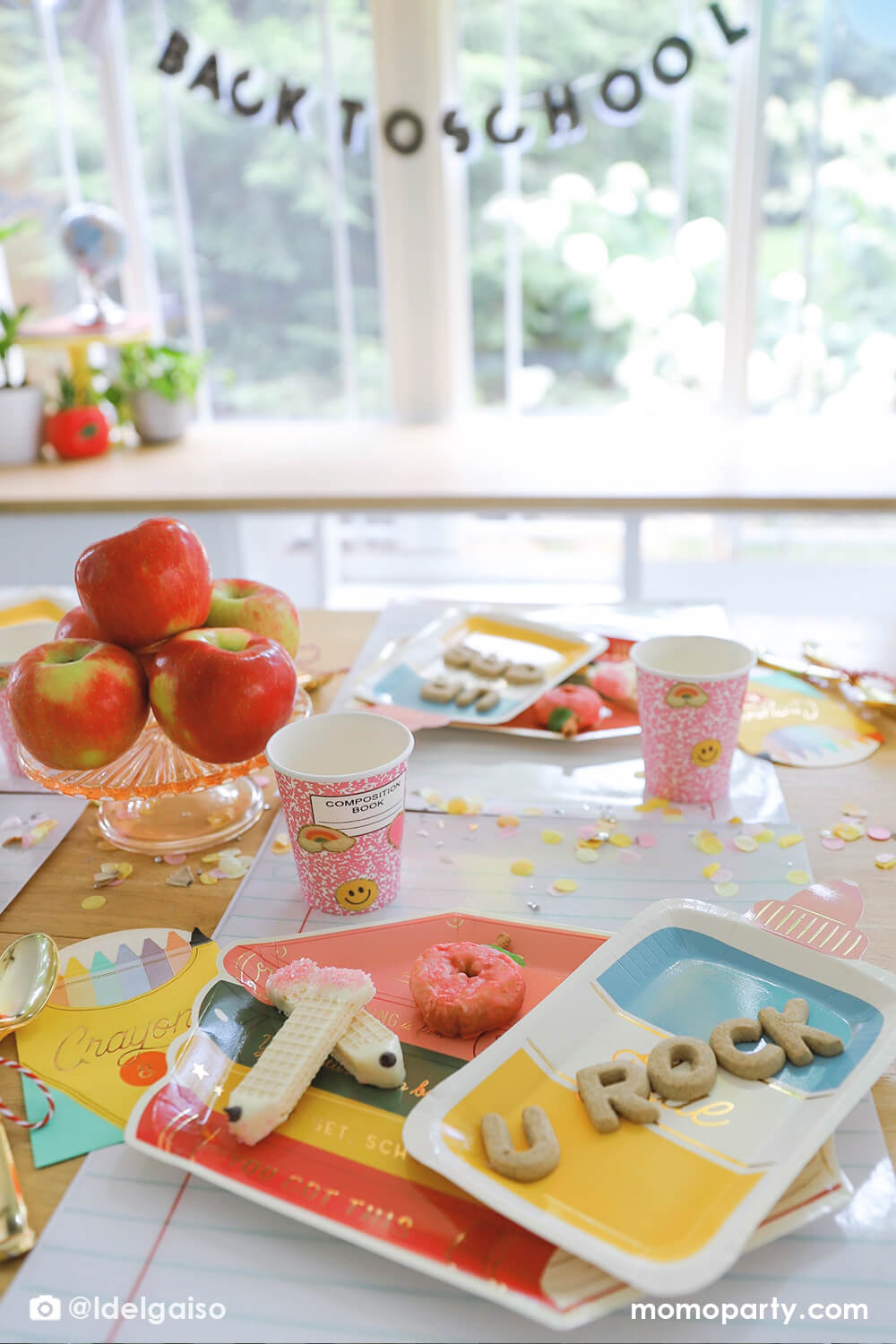 A close up shot of a festive back to school party table featuring multiple Momo Party's school themed party supplies including stack of books shaped plates, glue bottle shaped plates, crayon box shaped napkins, pink notebook party cups and giant notebook placemats. With pencil inspired confetti around and the stack of apples as the centerpiece on the table, plus the back to school banner hung above the table, this makes a perfect first day of school celebration for kids. 