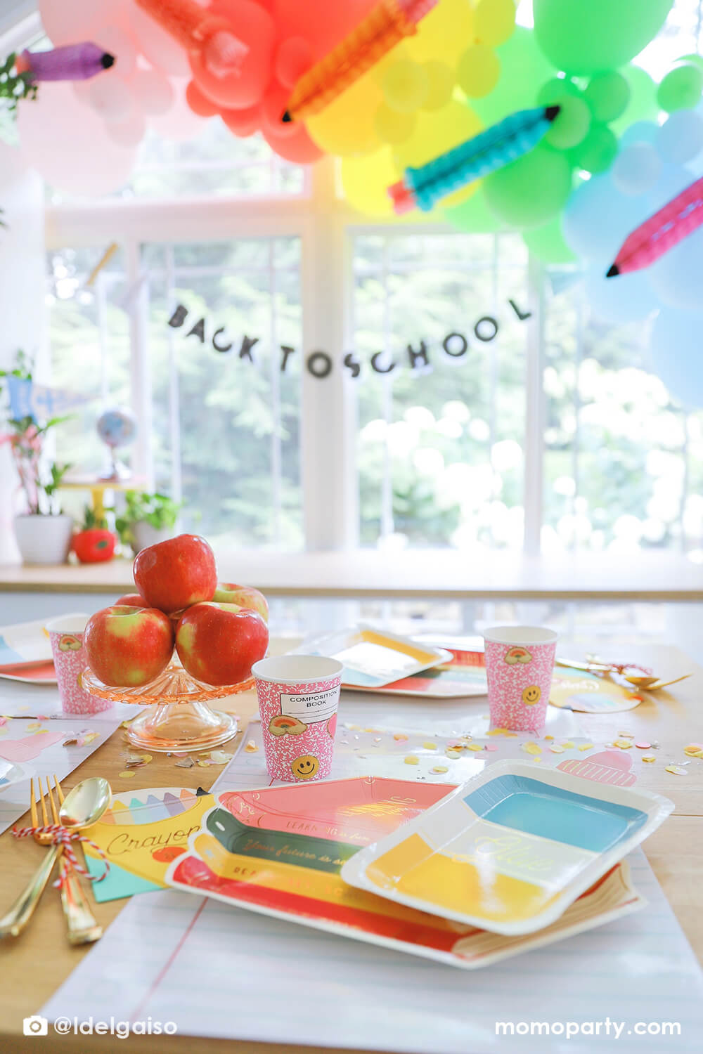 A festive back to school party featuring adorable school themed party supplies from Momo Party including the colorful stack of books shaped plates, glue bottle shaped plates,  crayon shaped napkins, pink notebook party cups, giant notebook placemats and pencil inspired confetti around. In the back there's a pastel balloon garland in rainbow colors adorned with matching pencil honeycomb, along with back to school banner hung underneath. Making this a perfect inspo for kid's first day of school celebration.