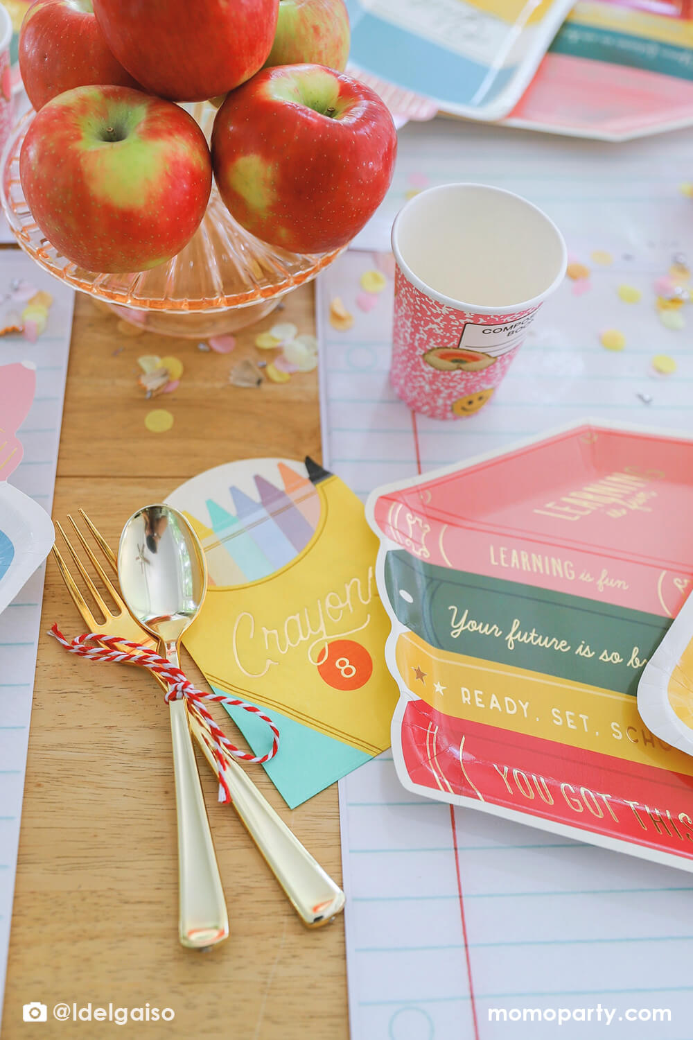 A closeup shot of Momo Party's crayon box shaped napkin and stack of books shaped plate on the giant notebook placemat from a back to school party table. Around plate is a pink notebook inspired party cup and some pencil inspired confetti around it. Along with the stack of apples in the middle of the table as the centerpiece, this simple table setting is perfect for kid's first day of school celebration.