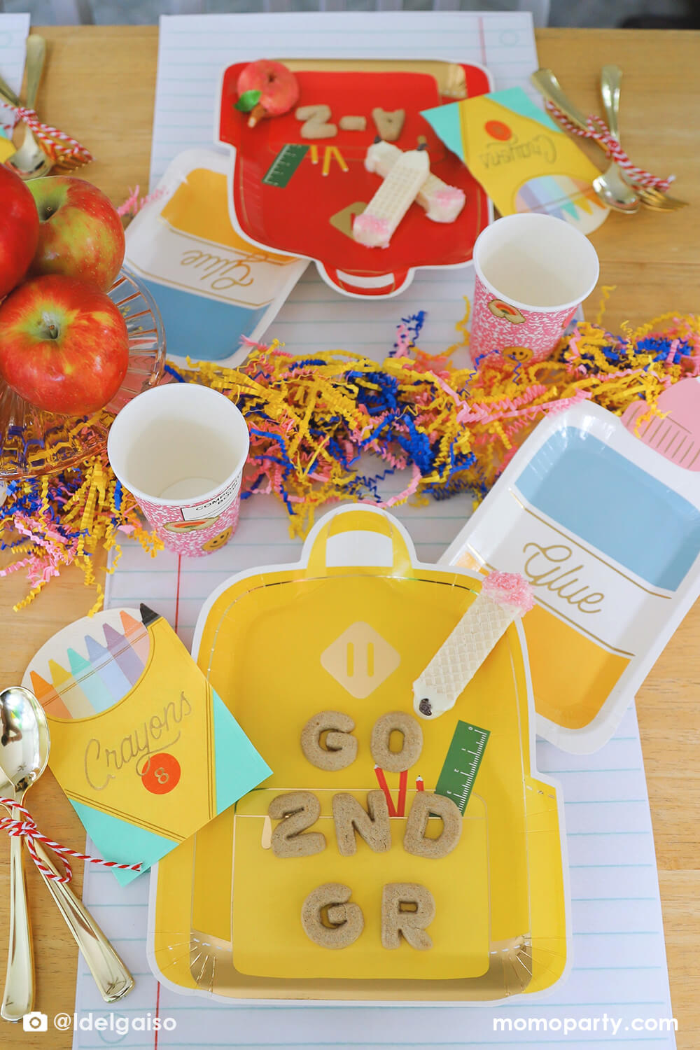 A festive back to school party tablescape featuring Momo Party's school themed party supplies including backpack shaped plate set in three different colors, glue bottle shaped plates, crayon box shaped napkins, pink notebook inspired party cups and giant notebook placemats. On the plates, there are alphabet shaped cookies spelling "Go 2nd Grader" and pencil shaped wafer cookies, with apples stacked as the centerpiece and some fun confetti, this makes a perfect first day of school celebration for kids.