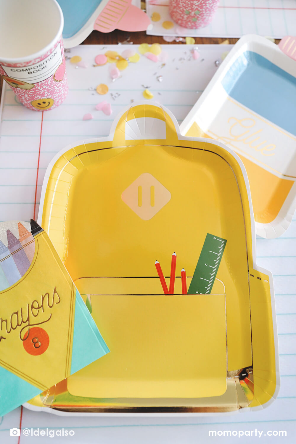 A close up shot on Momo Party's yellow backpack shaped plate by My Mind's Eye from a back to school party table featuring school themed party supplies including glue bottle shaped plates, pink notebook cups, crayon box shaped napkins and giant notebook placemats. With pencil inspired confetti, this makes a simple yet cute table setting for kid's first day of school celebration.