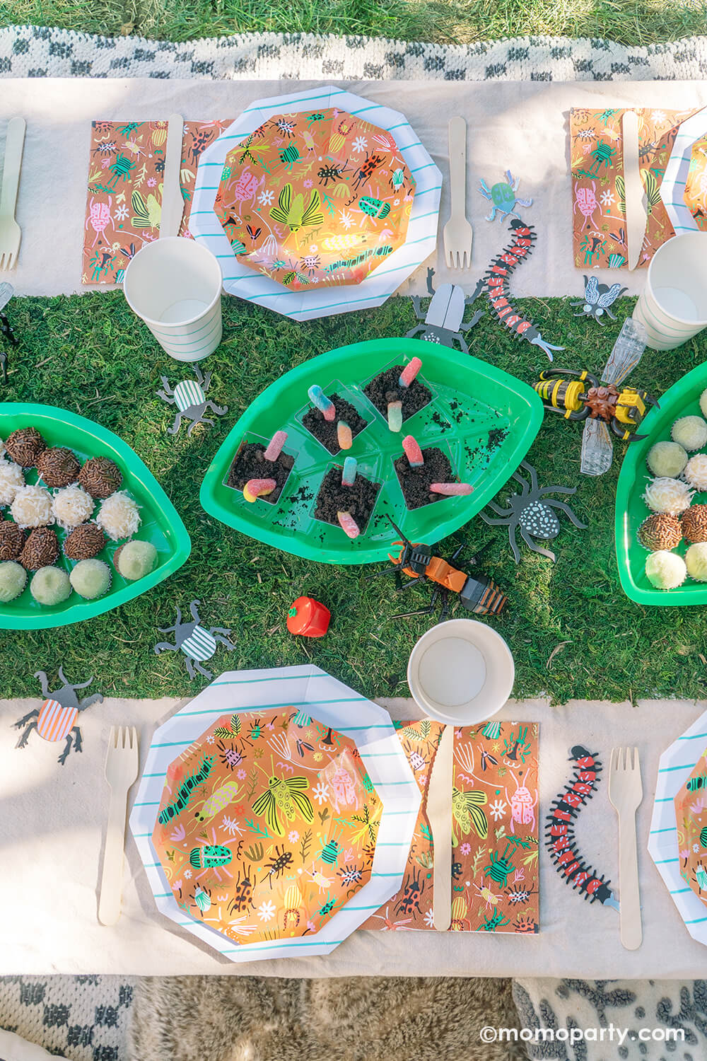 An insect bug themed birthday party table styled by Momo Party features backyard bug plates, napkins with aqua striped plates and party cups by Daydream Society. In the middle there's a faux grass table runner that was decorated with various lego insect toys and palm leaf shaped serving tray filled with insect themed treats and snacks including gummy worms, ladybug babybel cheeses and bug egg shaped brigadeiros, some playful and fun ideas for kid's insect themed bash.