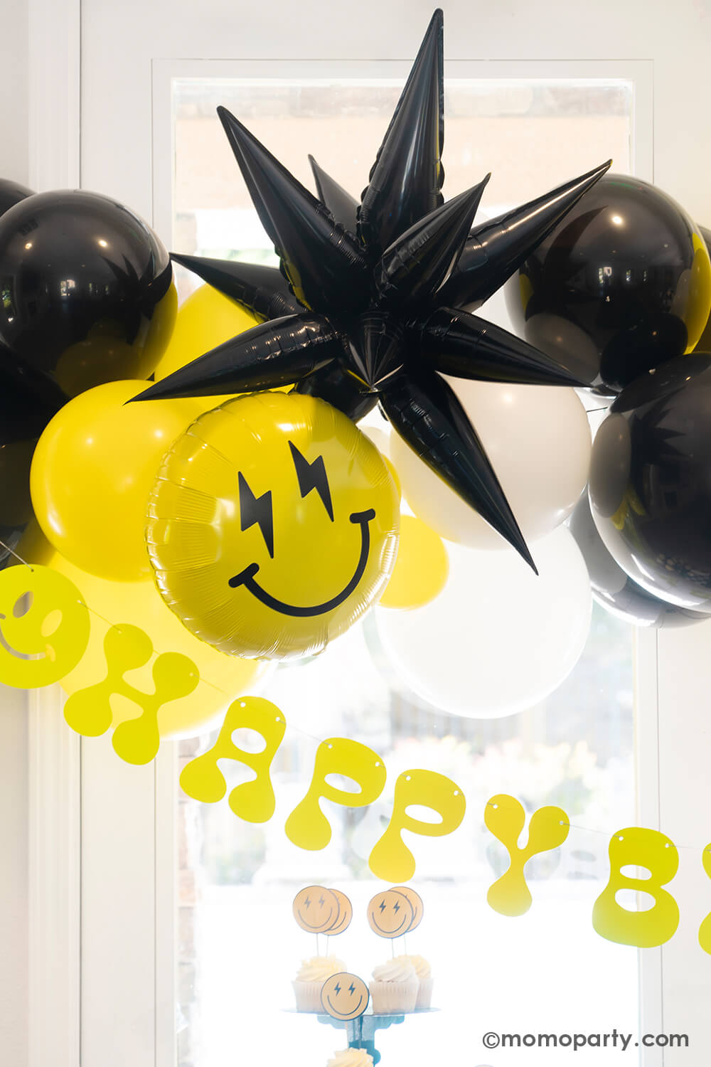 A close-up look at Momo Party's 'Cool Dude' themed Birthday Party. Featuring a captivating balloon garland in black, white, and yellow, accented by a black starburst foil balloon and a smiley face foil balloon with lightning bolt eyes. This stylish decor is complemented by a retro neon happy birthday banner. Perfect for a boy's 'One Happy Dude' themed first birthday, a 'Two Cool' themed second birthday party, 'Ten Rad Years' 10th birthday celebration, or kids birthday party for all ages. 