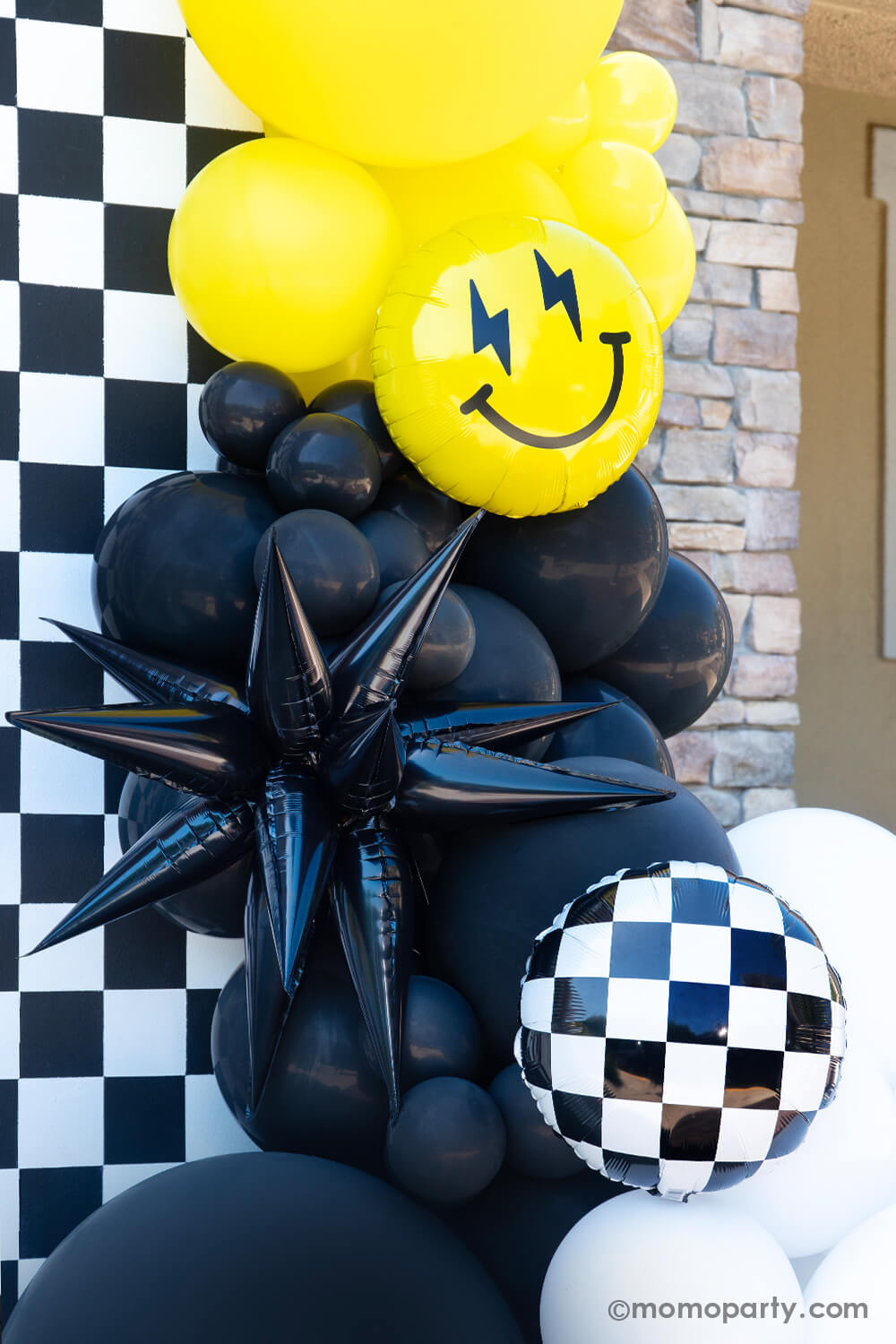 A close up look of Momo Party's "Cool Dude" themed Birthday Party, featuring  a balloon garlands in black, white and yellow adorned with a black starburst foil balloon,  Checkerboard Foil Balloon and smiley fact foil balloon with lightning bolt eyes, in front of the black and white checkerboard. This decoration is perfect for boy's "One Happy Dude" themed first birthday, a "Two Cool" themed second birthday party or "Ten Rad Years" 10th birthday party.  