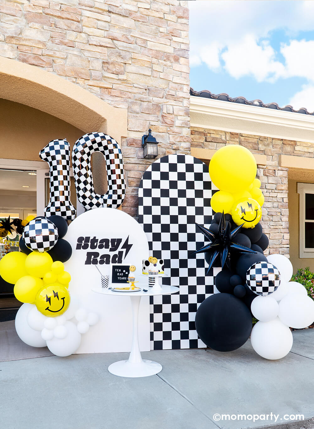 Momo Party's modern 'Cool Dude' 10th Birthday Party features a stylish balloon garland in black, white, and yellow, black starburst foil balloon, smiley face foil balloon, and Large Number 10 Checkered Foil Balloons. With a backdrop of b&w checkerboard and a 'Stay Rad' message, The table is set with Smiley Gold Foiled Paper Plates, Check It! Checkered Dinner Plates, Checkered Flag Party Cups, and Black Checkerboard Party Straws. Complete with a dotted fondant cake, it's a cool 'Ten Rad Years' celebration!