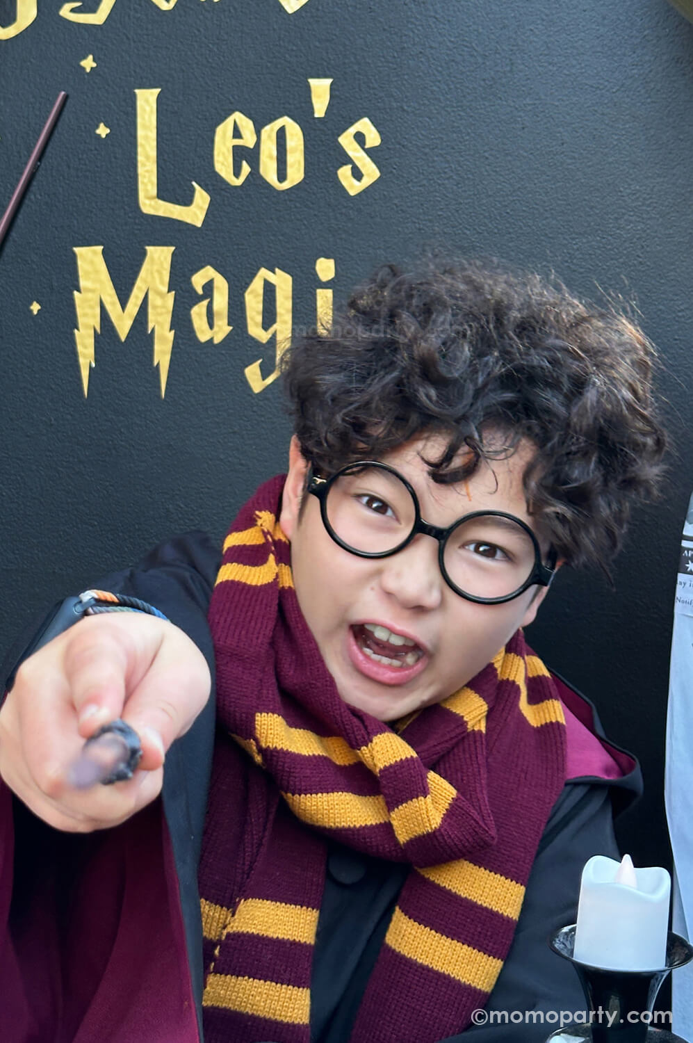 Momo Party hosted Harry Potter-themed birthday party featuring a boy dressed in a Gryffindor costume and wearing Harry Potter glasses available at momoparty.com. Adding authentic charm to the magical theme, the boy holds a wand while casting a spell in front of a black backdrop with the text '10 years of Leo's Magic,' creating a fully immersive Harry Potter experience.