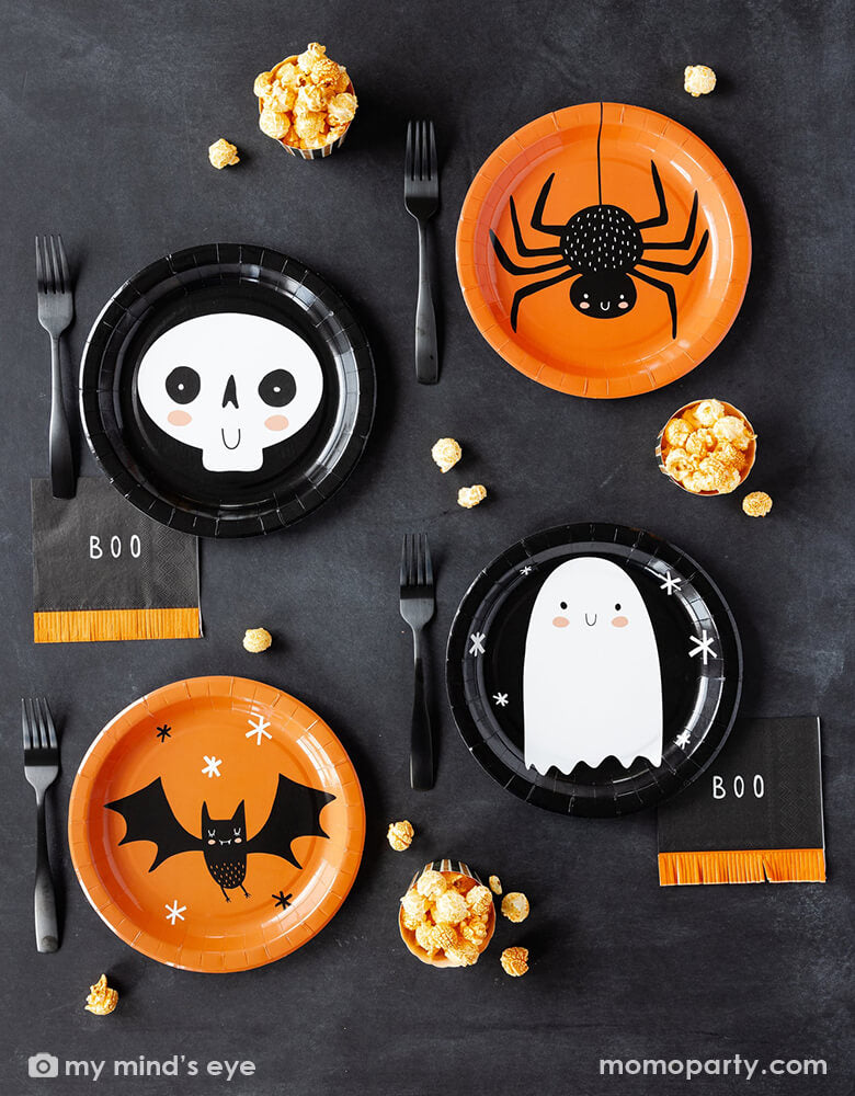 A kid's friendly Halloween party tablescape features Momo Party's classic Halloween party supplies including the friendly black and orange round plates with adorable character illustrations like a bat, a skull, a ghost and bat on them , the boo fringe small napkins, and the orange and black striped food cups filled with popcorn. This simple and modern place setting provide an effortless yet festive idea for celebrating Halloween with kids this spooky season.