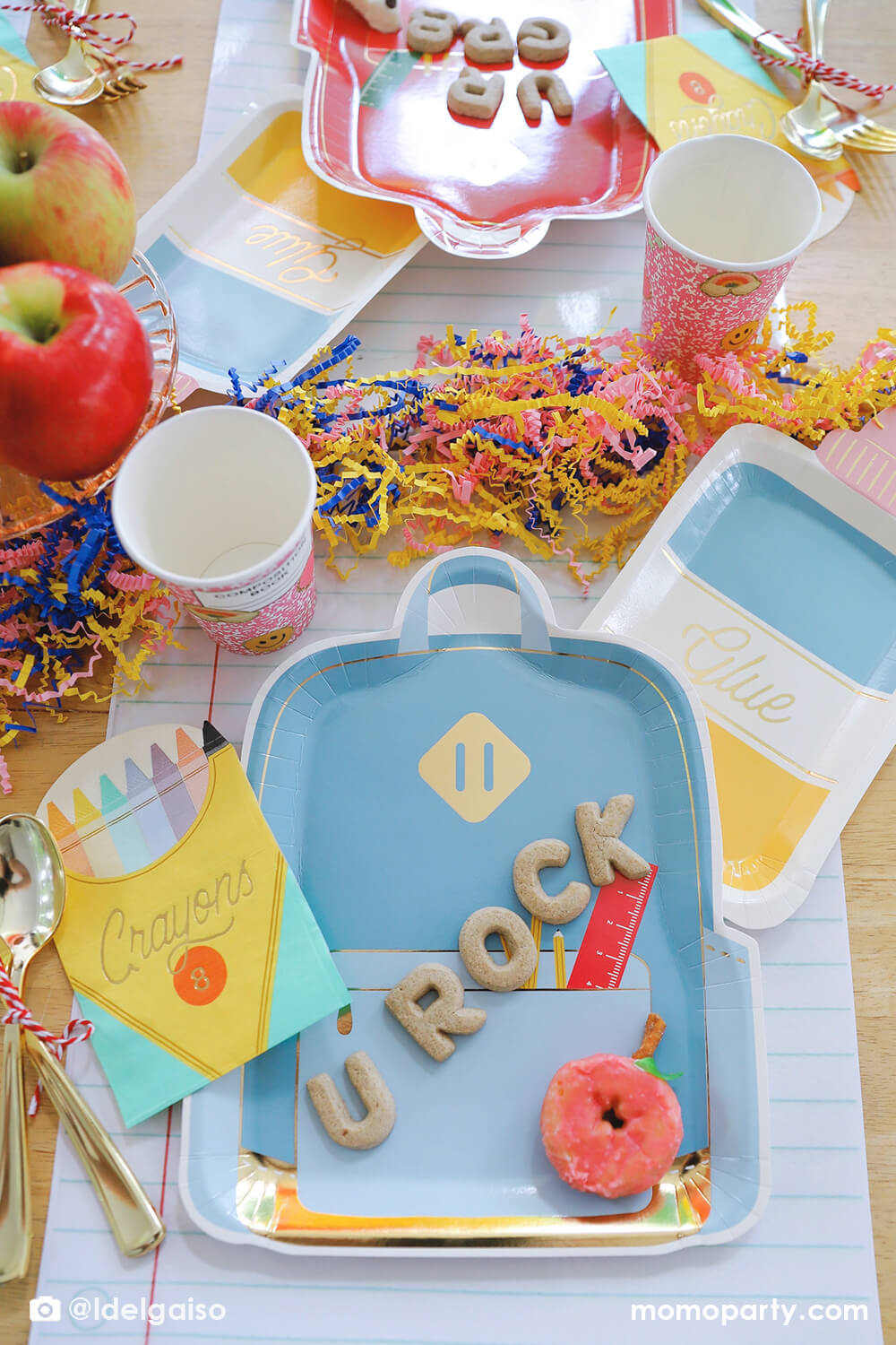 A festive back to school party tablescape featuring Momo Party's school themed party supplies including backpack shaped plate set in three different colors, glue bottle shaped plates, crayon box shaped napkins, pink notebook inspired party cups and giant notebook placemats. On the plates, there are alphabet shaped cookies spelling "U Rock" and apple shaped mini donuts, with apples stacked as the centerpiece and some fun confetti, this makes a perfect first day of school celebration for kids.