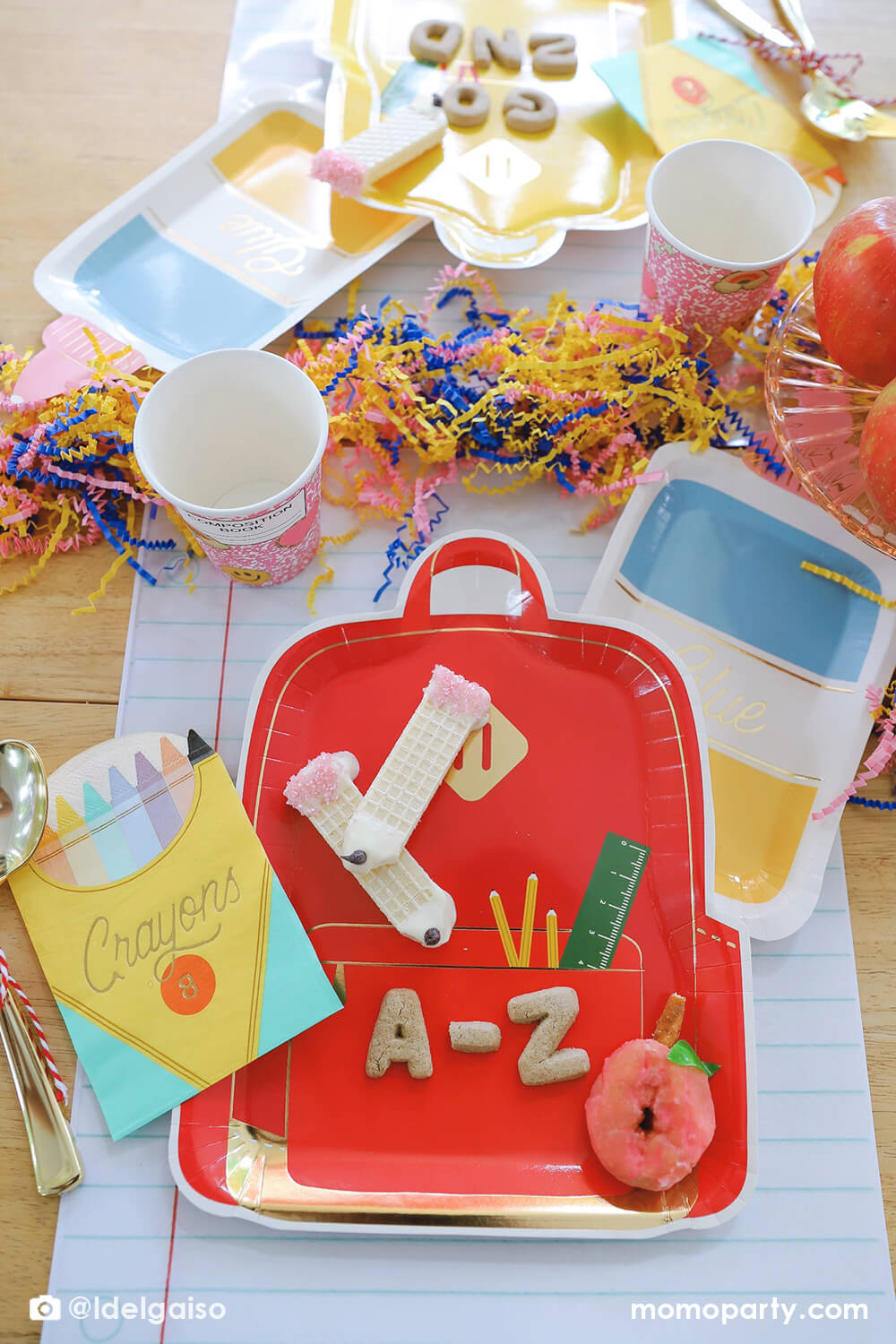 A festive back to school party tablescape featuring Momo Party's school themed party supplies including backpack shaped plate set in three different colors, glue bottle shaped plates, crayon box shaped napkins, pink notebook inspired party cups and giant notebook placemats. On the plates, there are alphabet shaped cookies and pencil shaped wafer cookies, with apples stacked as the centerpiece and some fun confetti, this makes a perfect first day of school celebration for kids.