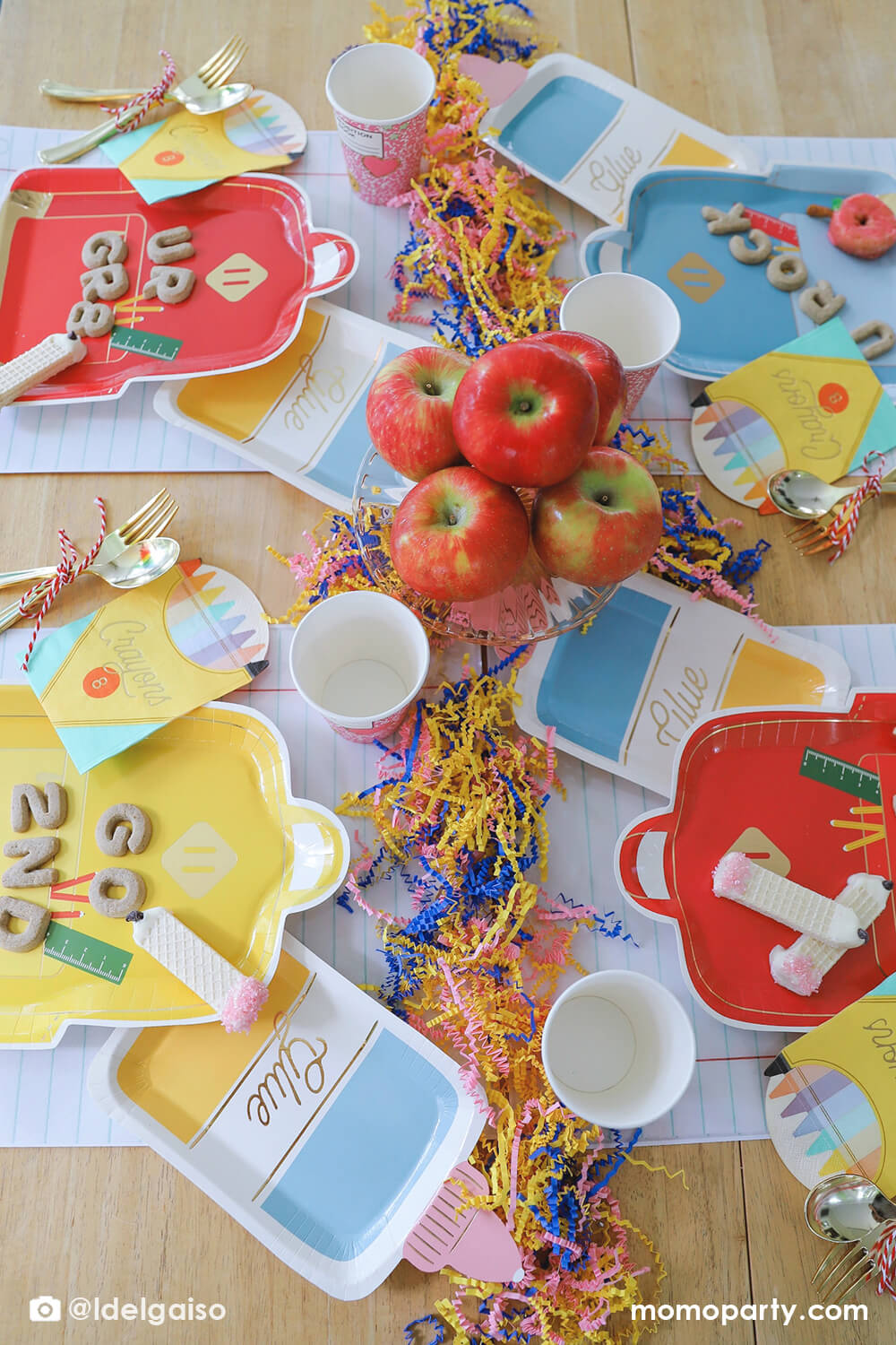 A overhead shot of a festive back to school party table featuring Momo Party's school themed party supplies including backpack shaped plate set in three different colors of red, blue and yellow, glue bottle shaped plates, crayon box shaped napkins, pink notebook party cups and giant notebook placemats. On the plates, there are alphabet shaped cookies and pencil shaped wafer cookies, with apples stacked as the centerpiece and some fun confetti, this makes a perfect first day of school celebration for kids.