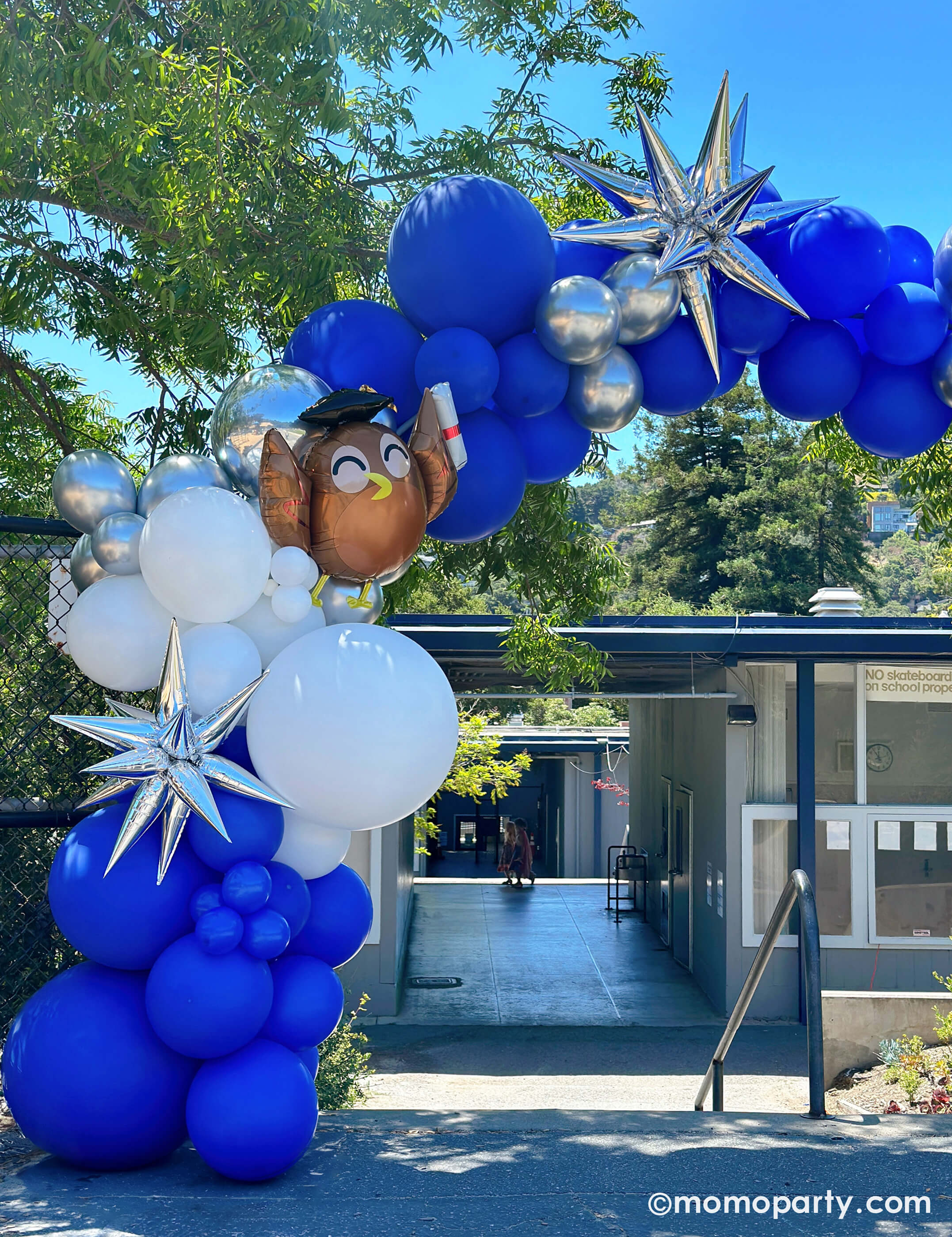 Elementary School Graduation Party by Momo Party. Featuring a stunning balloon arch in school colors of blue, white, and silver, accented with Graduate Owl Shaped Foil Balloons and a 12 Point Silver Starburst Foil Balloon in front of the school staircase. This arch is a big hit, serving as a graduation gate for students to pass through and a photo backdrop to capture last day of school memories.