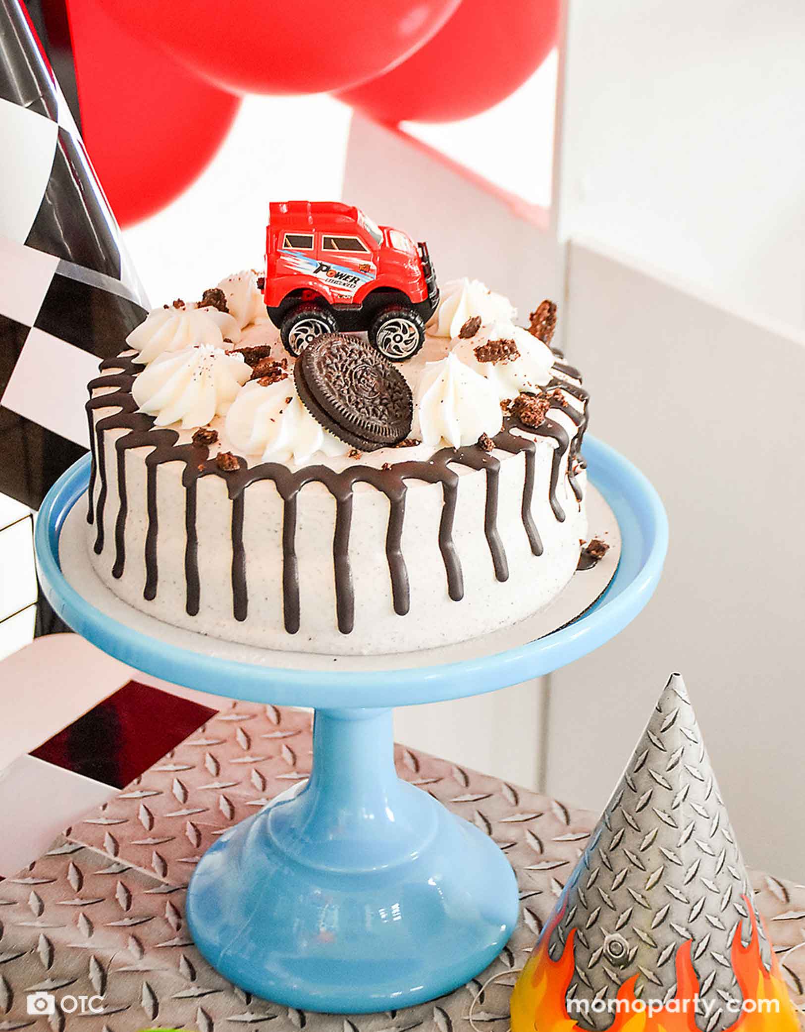 A monster truck themed birthday party table with a Oreo ice cream birthday cake decorated with Momo Party's red pull-back toy monster truck toy. On the table there's a party hat and a tablecloth in the steel texture design, along with race car checkered flags and red balloons, a fun and exciting monster truck birthday party awaits!