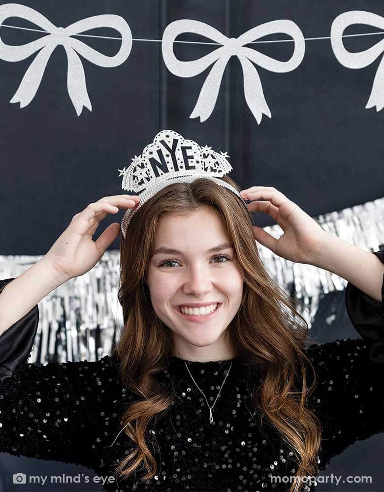 A young lady in a black sequin top wearing Momo Party's NYE silver glittered crown headband by My Mind's Eye. In her back hung a silver tinsel party banner and a silver glittered bow party garland, making this a modern and chic New Year's Eve countdown party decoration inspiration for this festive Holiday season. 