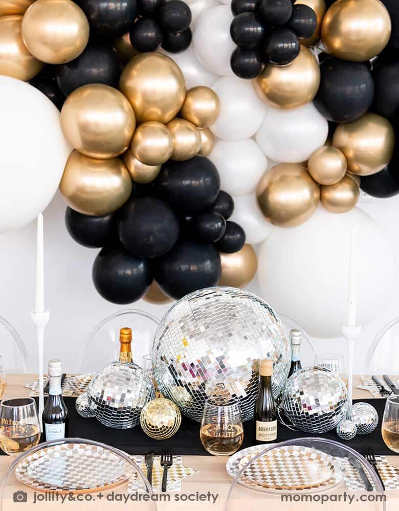 A gorgeous New-Year's-Eve-Party-Table set up featuring a balloon garland of black, gold and silver balloons, table set with Momo Party's silver and gold checkered tableware including plates, napkins and cups, in the middle there are some disco ball decorations in different sizes as the centerpiece, making this a chic and glimmer look for New Year's Eve countdown party. 