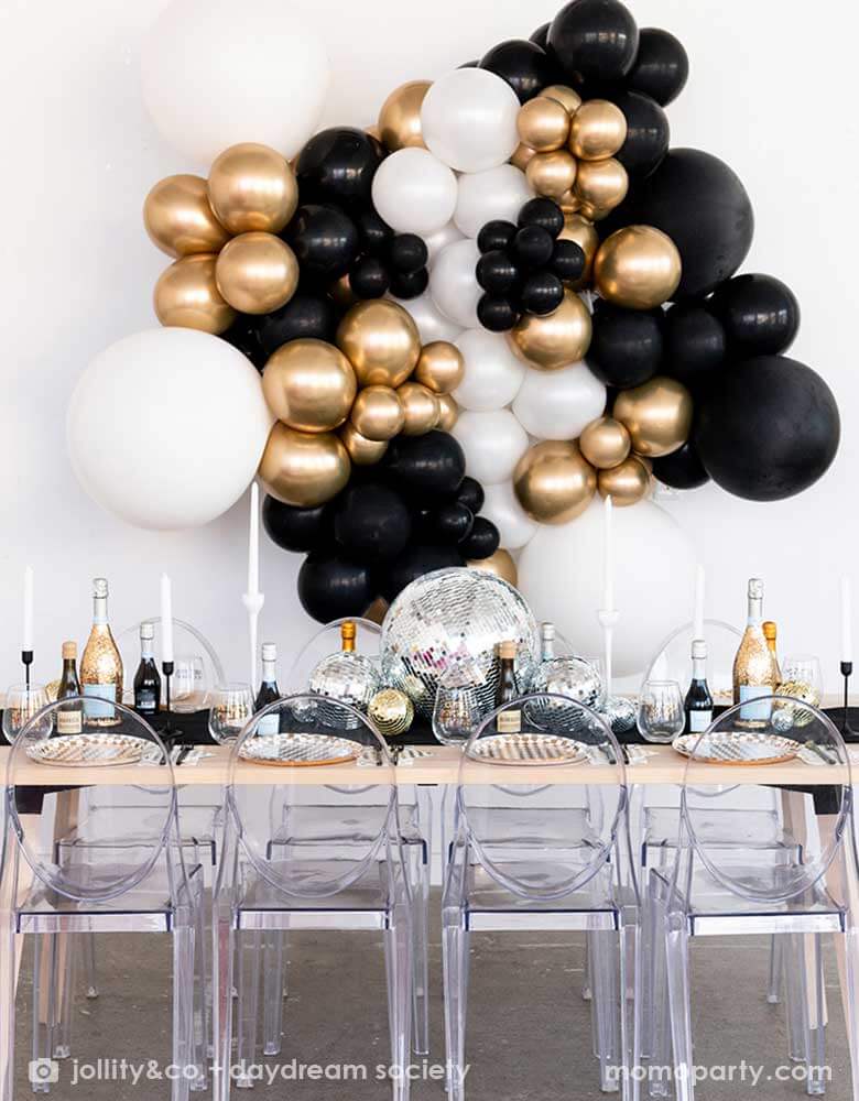 A gorgeous New-Year's-Eve-Party-Table set up featuring a balloon garland of black, gold and silver balloons, table set with Momo Party's silver and gold checkered tableware including plates, napkins and cups, in the middle there are some disco ball decorations in different sizes as the centerpiece, making this a chic and glimmer look for New Year's Eve countdown party. 