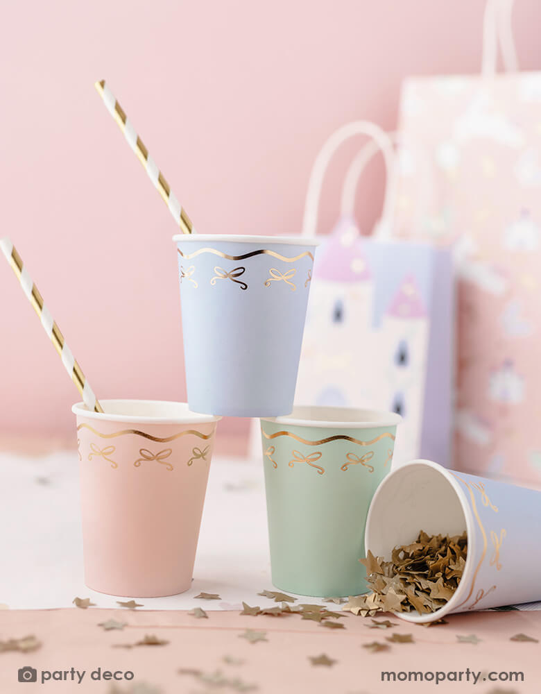 A pink party table featuring Momo Party's 7.4 oz pastel bow party cups in 3 different pastel colors with gold striped straws by Party Deco. Around the cups, there's some gold star confetti spread across the table. In the back there are some girl's party bags. 