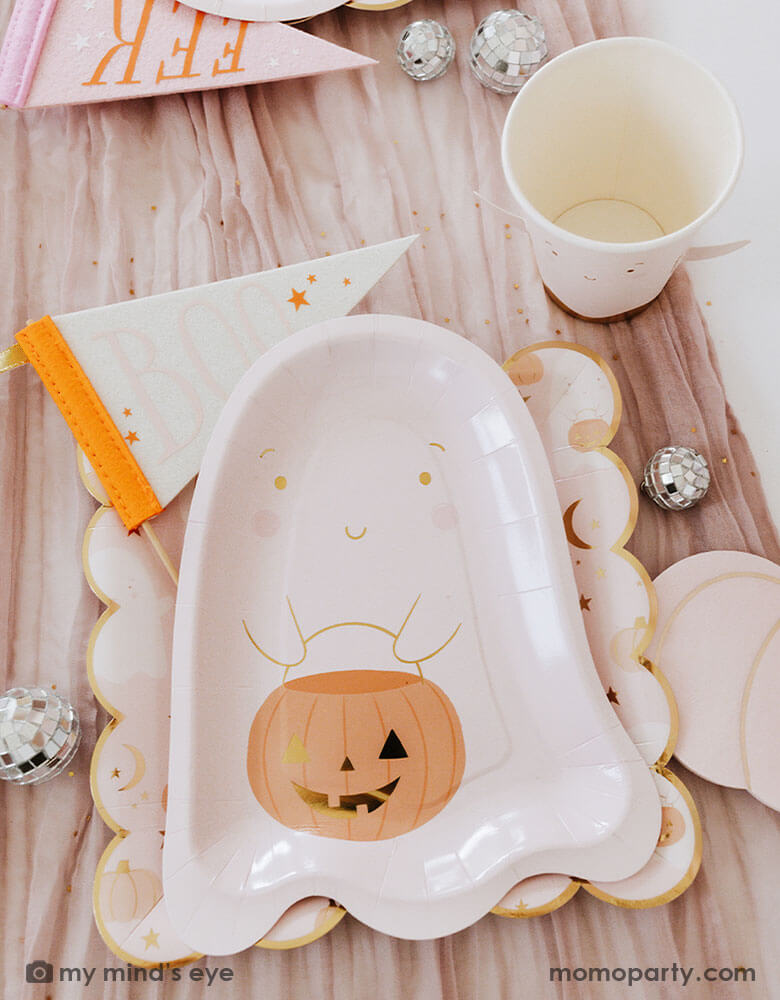 A Halloween party table filled with pink Halloween ghost themed party supplies from Momo Party including the pink ghost shaped paper plates with a jack-o-lantern, the 10" x 10" pink ghost scattered scalloped dinner plates, ghost party cup and Boo and EEK felt party pennants. With mini disco ball decorations, it makes a great inspo for a kid's friendly pink Halloween party this spooky season!