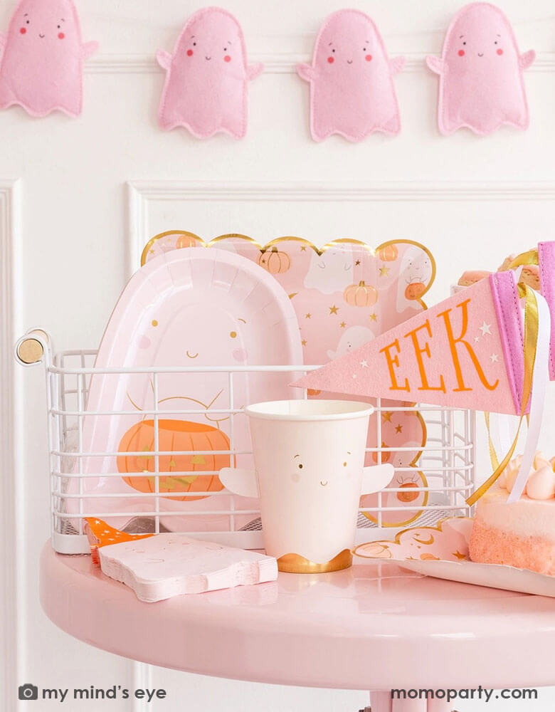 A pink Halloween party table featuring adorable party supplies from Momo Party including the 12 oz pink ghost shaped party cup with adhesive arm attachment, the ghost scattered scallop dinner plates, some ghost shaped napkins, a pink ghost shaped die cut plate with a smiley ghost holding a jack-o-lantern. Along with the pink felt party pennant which says "EEEK" on it and the pink felt ghost banner hung on the wall behind the table, this makes a great look for kid's not-so-spooky Halloween party.