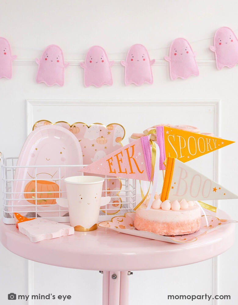 A pink Halloween party table featuring multiple adorable ghost themed party supplies from Momo Party including the 12 oz ghost shaped party cup, the ghost scattered scallop-edged dinner plates, ghost shaped napkins, a pink ghost shaped die cut plate with the ghost holding a jack-o-lantern. Along with a pink cake topped with felt pennants that have "SPOOKY," "EEK" and "BOO" on them, plus the pink felt ghost banner hung on the wall behind. Perfect for a kid's friendly Halloween bash!