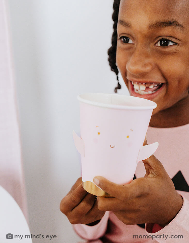 A school age girl holding Momo Party's 12 oz Ghost Paper Party Cup by Occasions By Shakira. An adorable and cute party cup for a kid's friendly pink Halloween celebration in the spooky season.