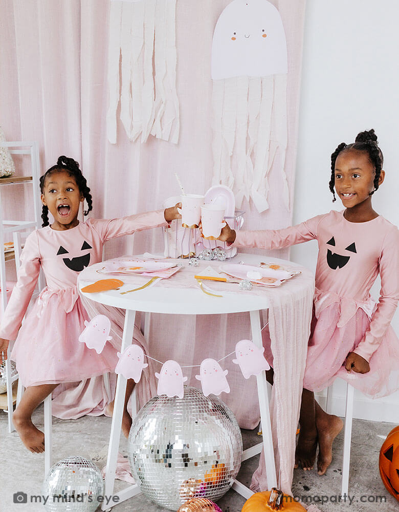 Two school aged girls in pink shirts that have Jack-o-lantern design on them and pink tutu sitting at a party table holding Momo Party's ghost shaped party cups. On the party table there are multiple pink Halloween party tableware spread out and the side is adorn with the pink ghost felt banner. Under the table there are some big disco balls and in the back hung some ghost paper decorations with streamer - a sweet and cute decoration for a Halloween celebration that is kid's friendly.