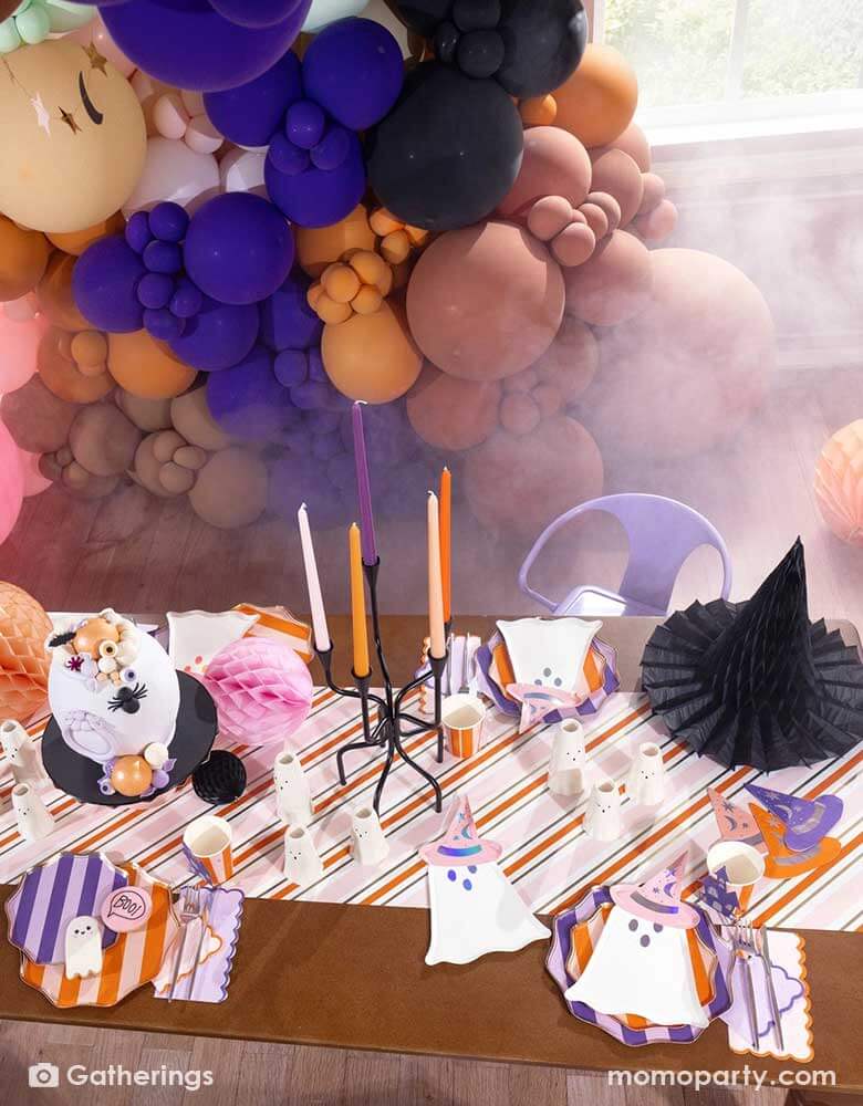 A festive pink Halloween party table set up featuring Momo Party's Bewitching ghost shaped plates, purple striped dinner plates, bewitching witch shaped hat napkins in three different colors of purple, orange and peach laid on a pink, orange and gold striped table runner. With some honeycomb pumpkins and witch hat decorations as the centerpiece, plus a whole of balloon garland in the colors of orange, brown, purple, making this an great inspo for a chic and cute Halloween party setup for this spooky season!