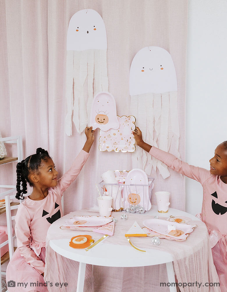 Two school aged girls in pink shirts that have Jack-o-lantern design on them and pink tutu sitting at a party table holding Momo Party's ghost shaped party plates. On the party table there are multiple pink Halloween party tableware spread out and the side is adorn with the pink ghost felt banner. Under the table there are some big disco balls and in the back hung some ghost paper decorations with streamer - a sweet and cute decoration for a Halloween celebration that is kid's friendly.