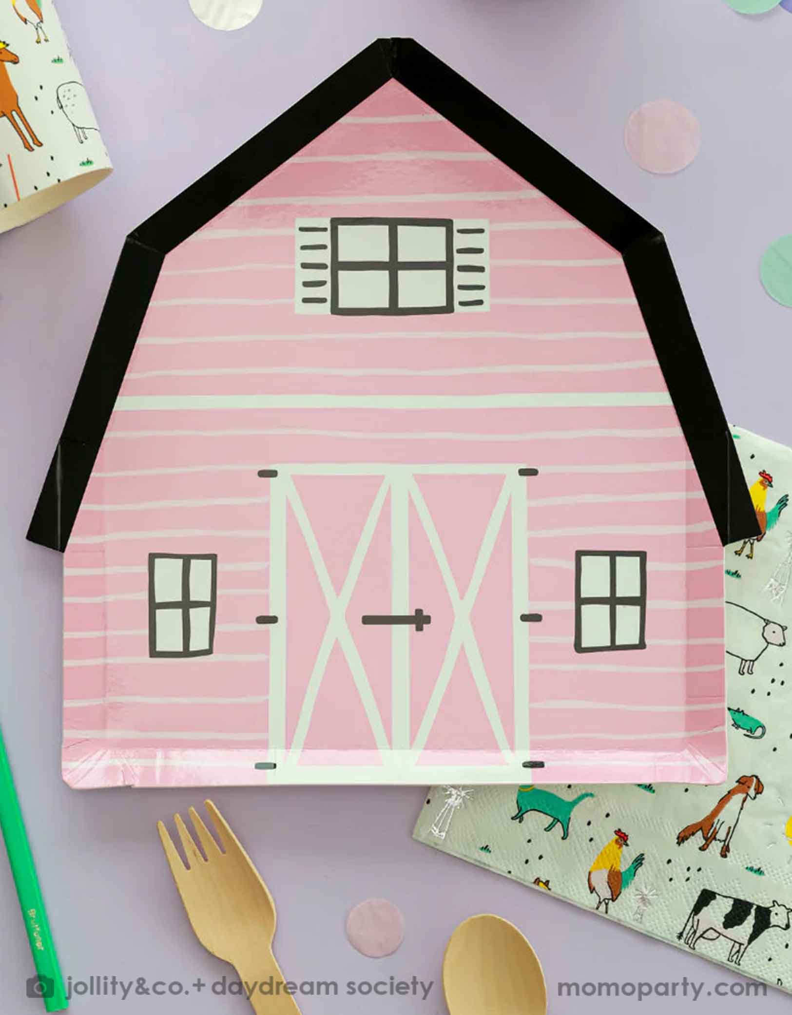 A kid's farm themed birthday party tablescape featuring Momo Party's pink on the farm barnhouse shaped plate and farm motif large napkins and party cups with farm animals including cows, chickens, dogs, pigs etc, with pink confetti and wooden cutlery around.