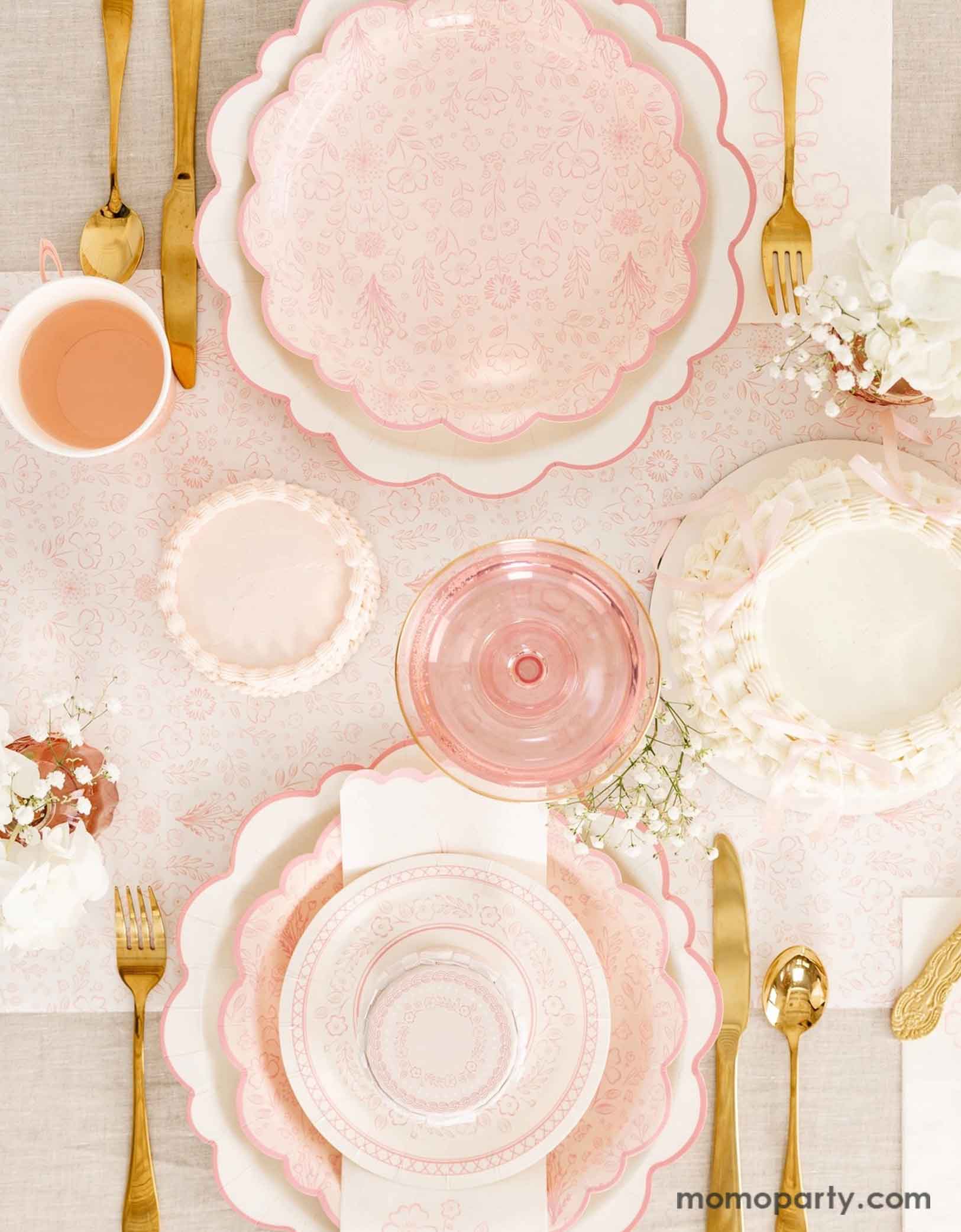 An elegant tablescape featuring Momo Party's Pink Pembroke tableware collection including the 12" Pembroke Pink Edge Scallop Plates paired, 9" Pembroke Pink floral Plates, 7" Pembroke Pink Plates, Pembroke Pink napkins and party cups. Underneath the plates is the Pembroke Pink floral paper table runner. With the gold foil utensil set, a pretty floral arrangement and pastel pink butter cakes and glasses of rose champagne, this table setting is perfect for an Easter celebration or Mother's Day party.