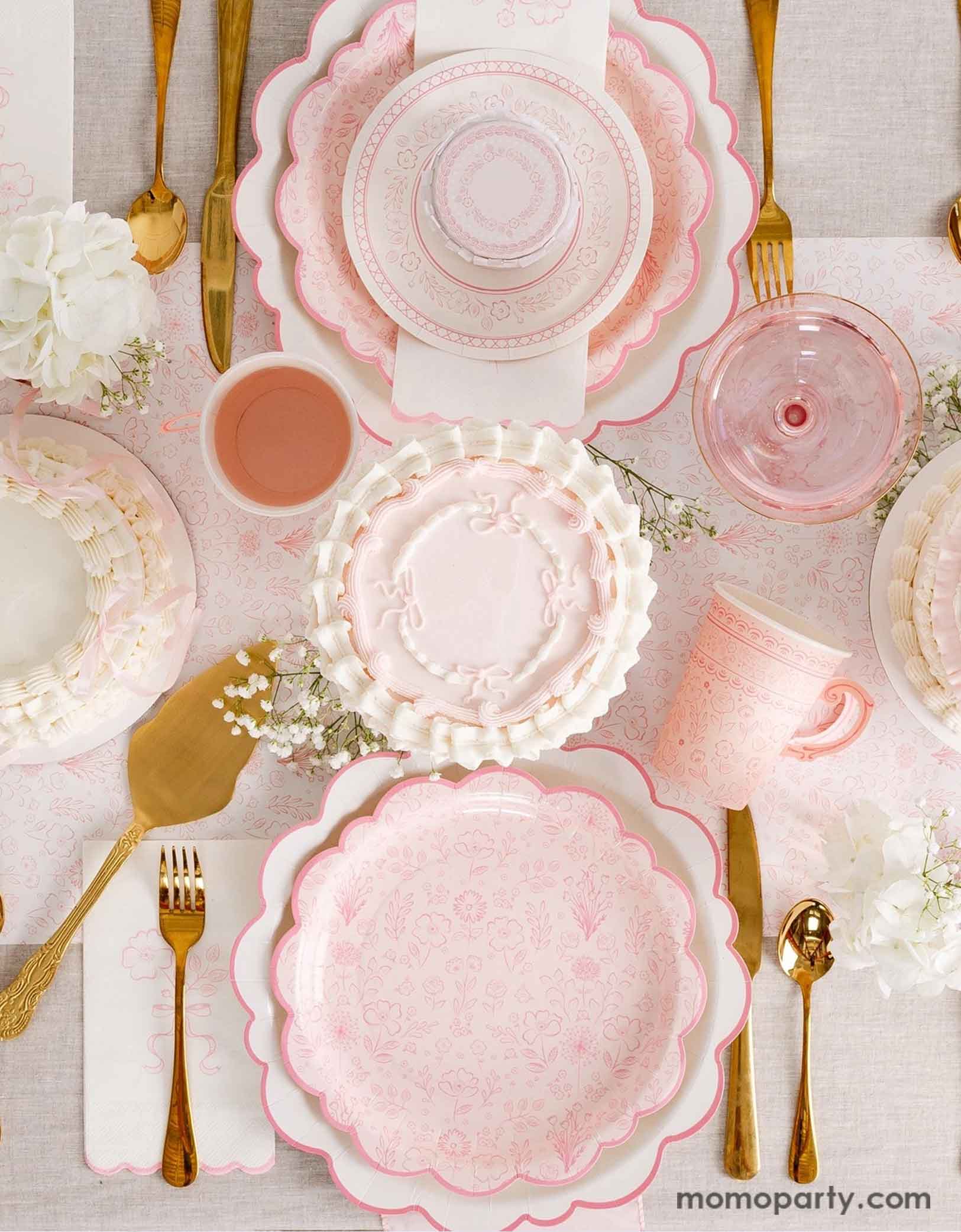 An elegant tablescape featuring Momo Party's Pink Pembroke tableware collection including the 12" Pembroke Pink Edge Scallop Plates paired, 9" Pembroke Pink floral Plates, 7" Pembroke Pink Plates, Pembroke Pink napkins and party cups. Underneath the plates is the Pembroke Pink floral paper table runner. With the gold foil utensil set, a pretty floral arrangement and pastel pink butter cakes and glasses of rose champagne, this table setting is perfect for a spring Easter inspired celebration.