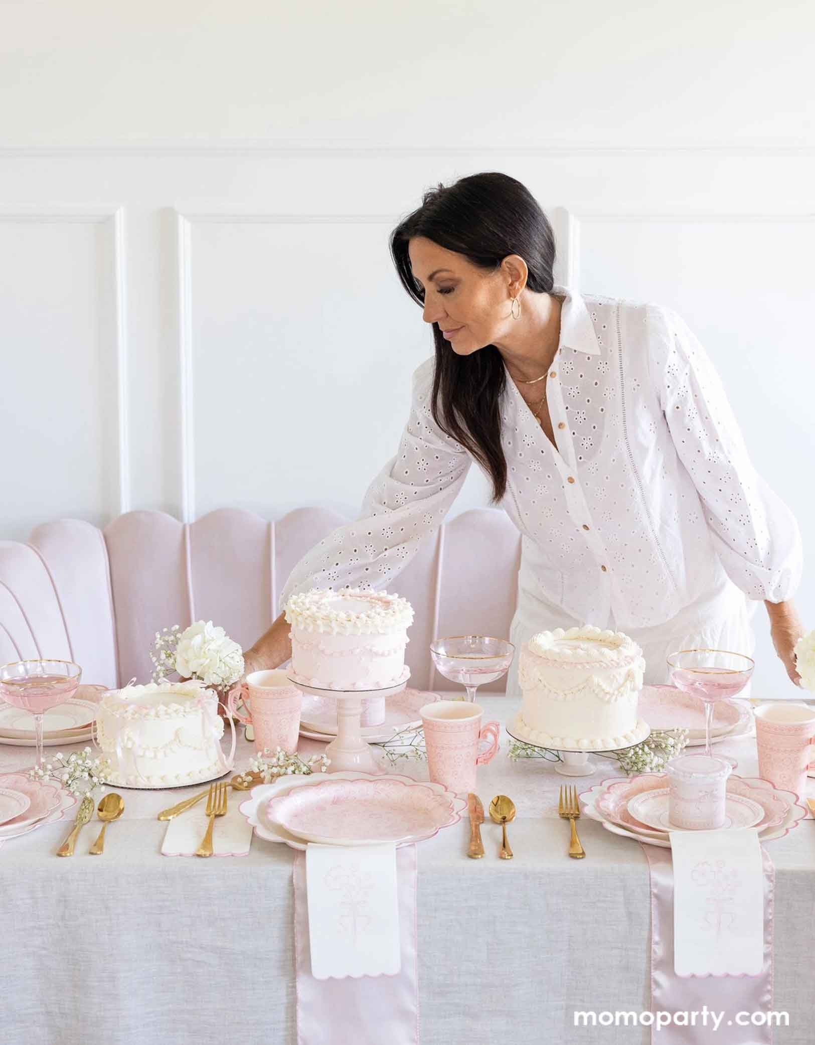 A lady is setting up an elegant party table featuring Momo Party's Pink Pembroke tableware collection including the 12" Pembroke Pink Edge Scallop Plate paired, 9" Pembroke Pink floral Plate, 7" Pembroke Pink Plate, the Pembroke Pink napkin and the matching party cup. With pretty floral arrangements, pink buttercream cakes and pink champagne glasses, this table setting is perfect for a spring inspired celebration, be it an Easter celebration, a bridal shower, a tea party birthday bash or Mother's Day party.