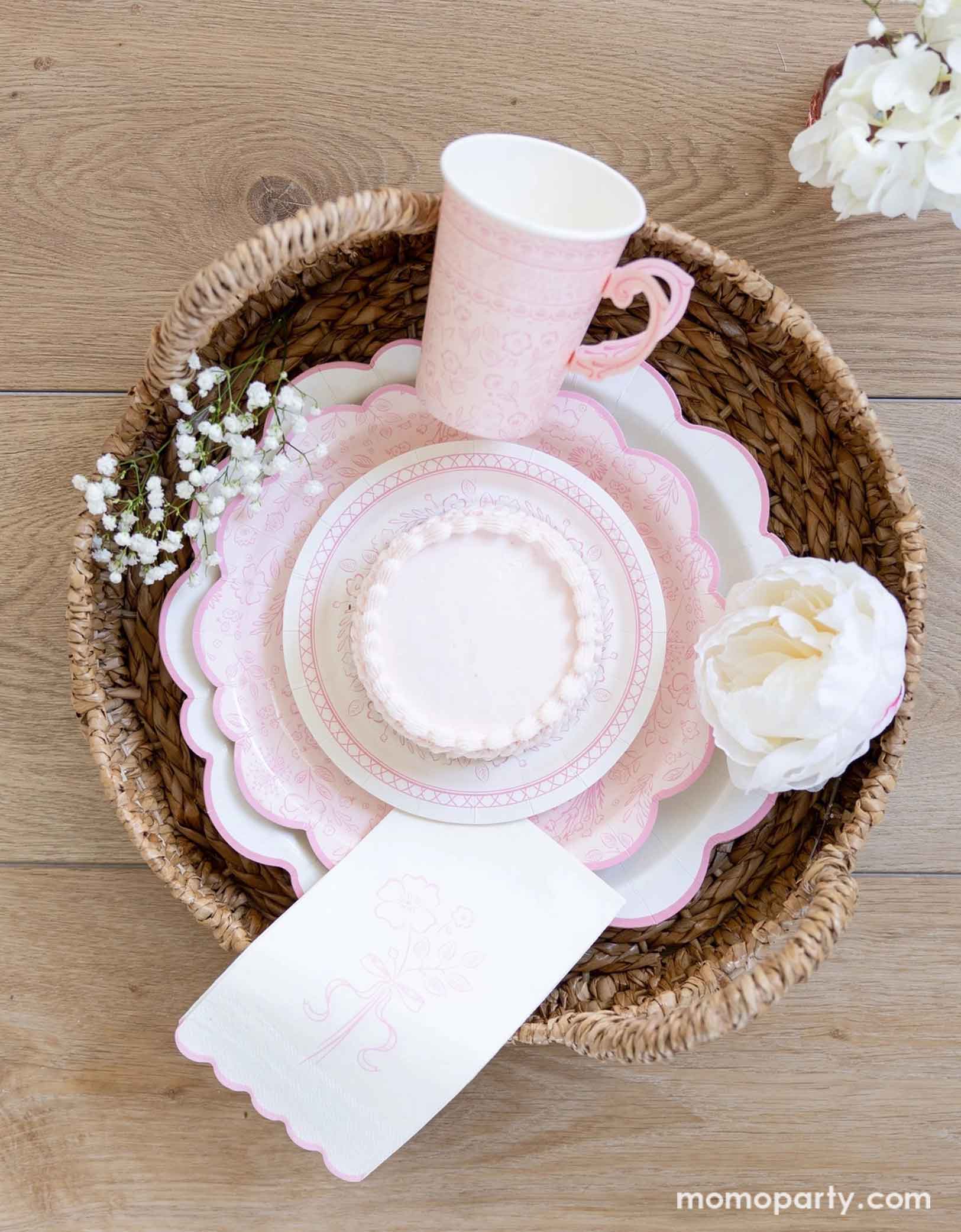 A woven basket filled with Momo Party's Pink Pembroke tableware collection including the 12" Pembroke Pink Edge Scallop Plate paired with 9" Pembroke Pink floral Plate and Pembroke Pink Edge Scallop Napkin. Underneath the plates is the Pembroke Pink floral paper table runner. With the gold foil utensil set, a pretty floral arrangement and pastel pink butter cakes and a glass of rose champagne,.