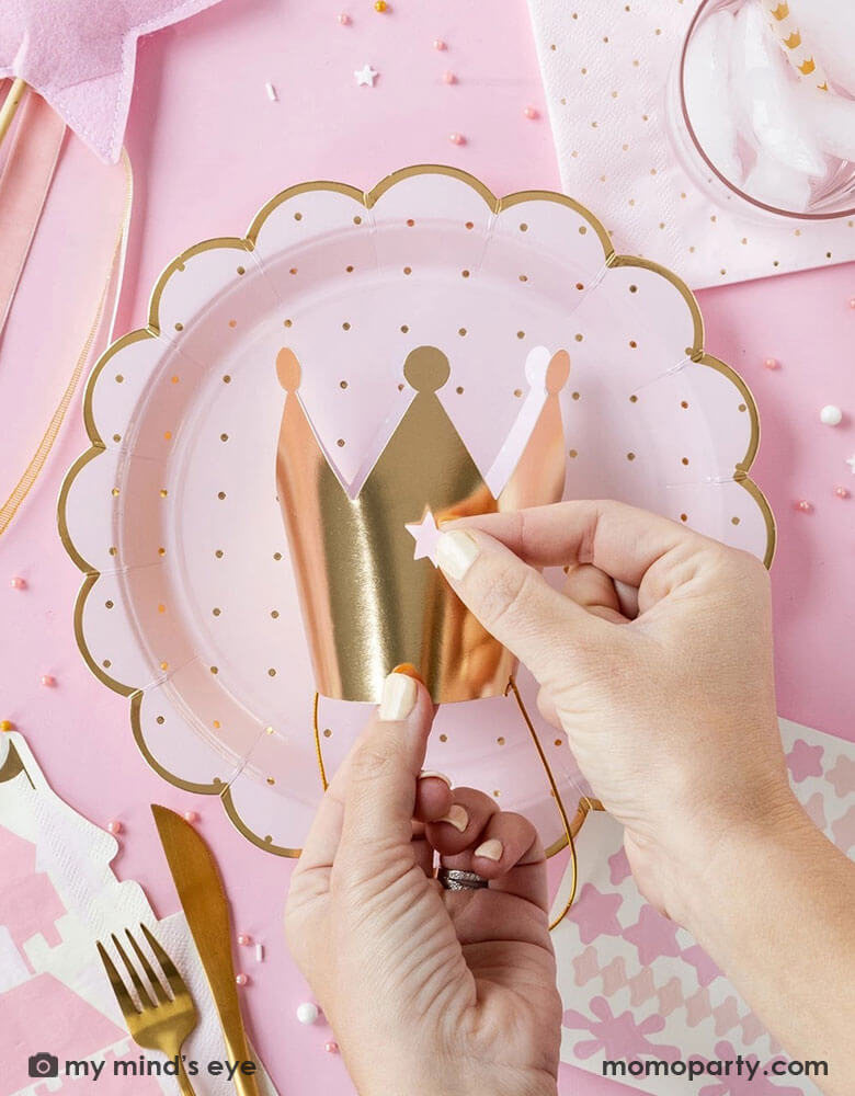 A girl's princess themed party table featuring Momo Party's pink princess themed party supplies including scalloped plates with gold polka dots, napkins, a pink star shaped wand and a gold paper crown being decorated with stickers, making this a great inspo for girl's princess themed birthday party celebration.